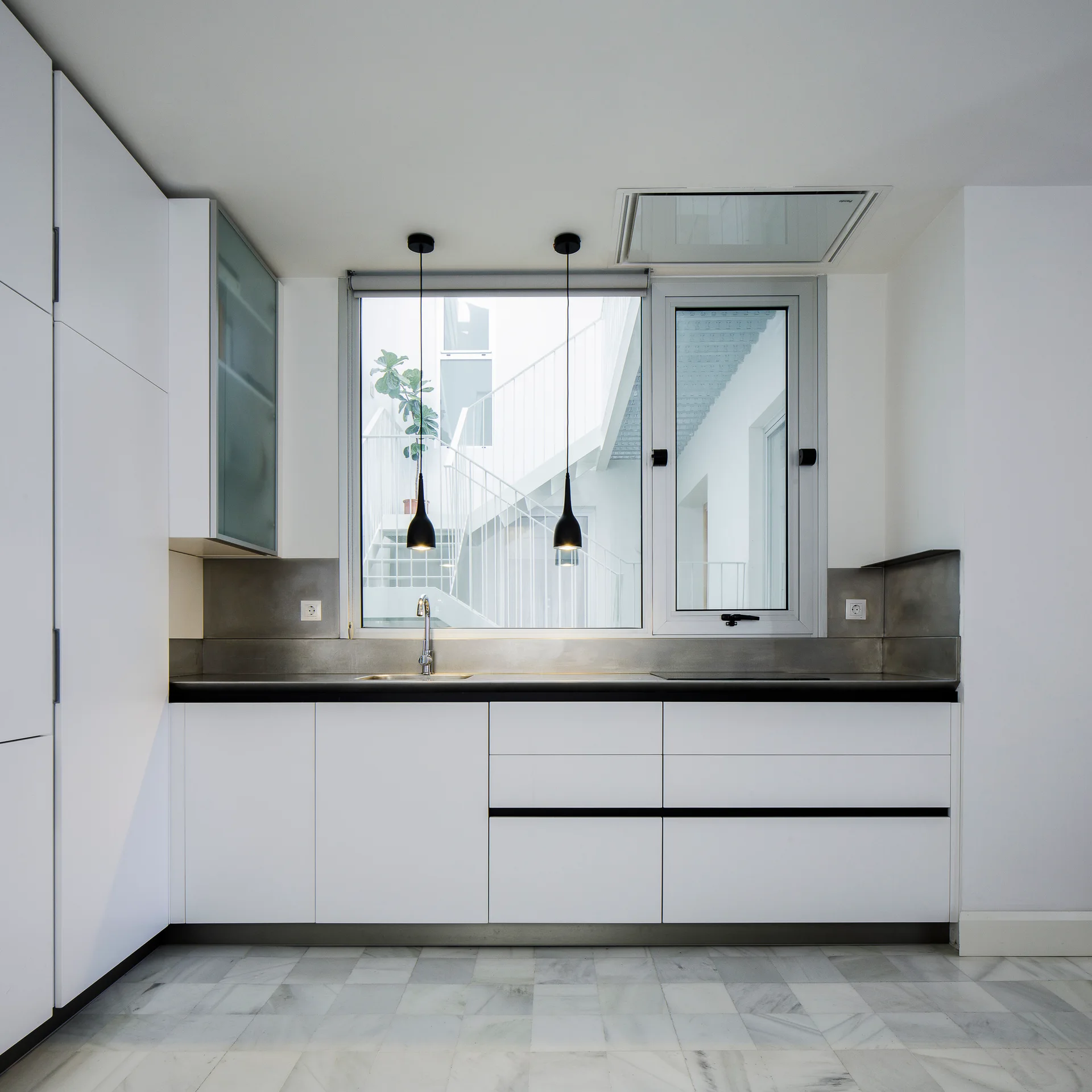 A minimalist white kitchen featuring handleless cabinets, a dark countertop, a brushed metal backsplash, and a large window overlooking an outdoor staircase.