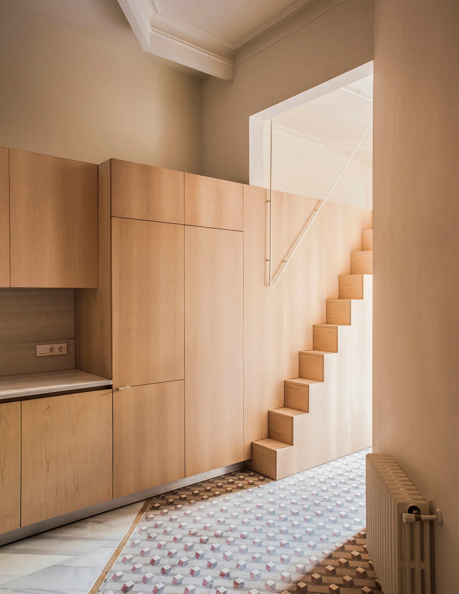 Minimalist light wood kitchen cabinets integrate seamlessly with a wooden staircase above geometric patterned tile flooring.