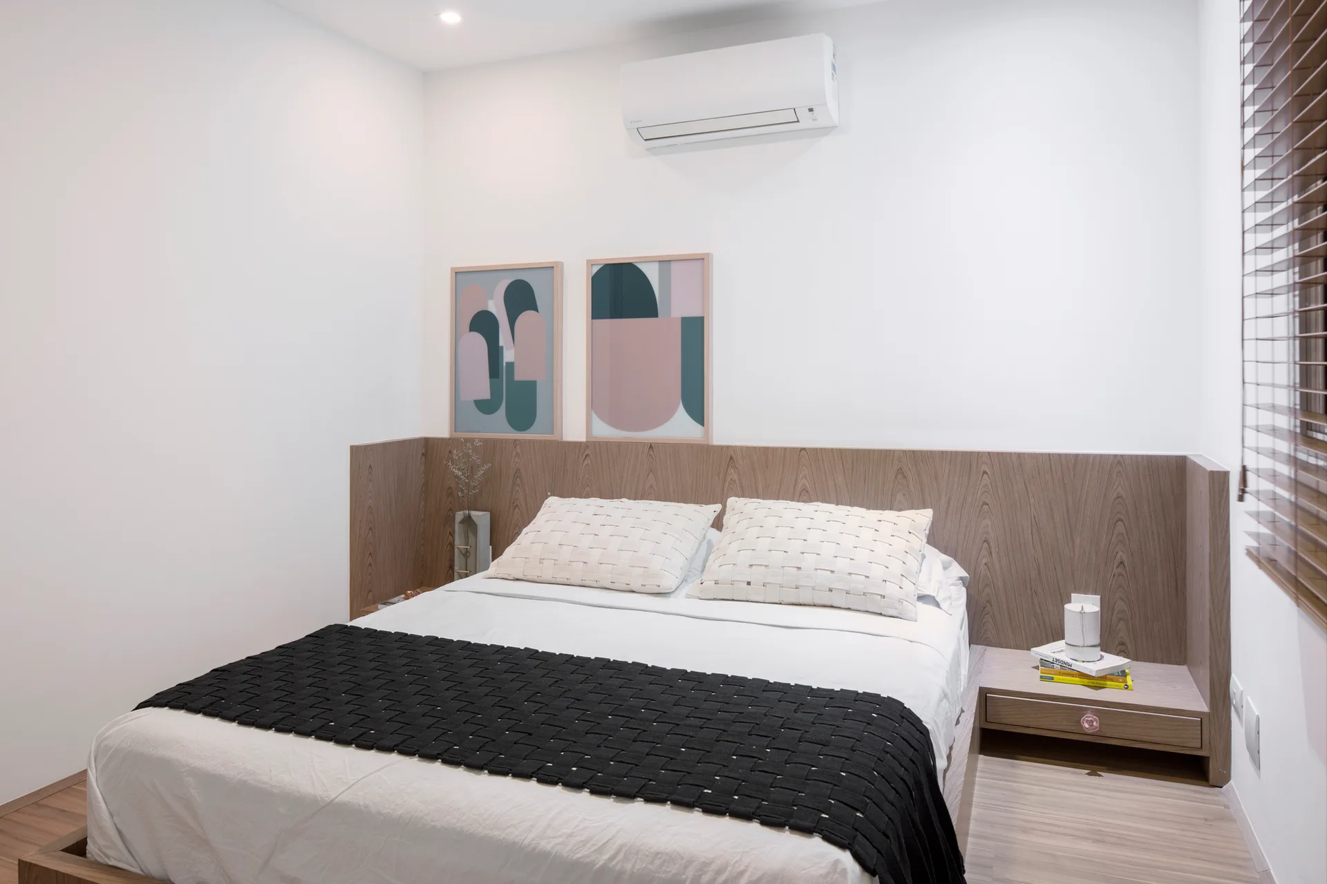 A modern bedroom features a bed with white linens and a black runner, set against an integrated wood headboard with abstract framed art above.