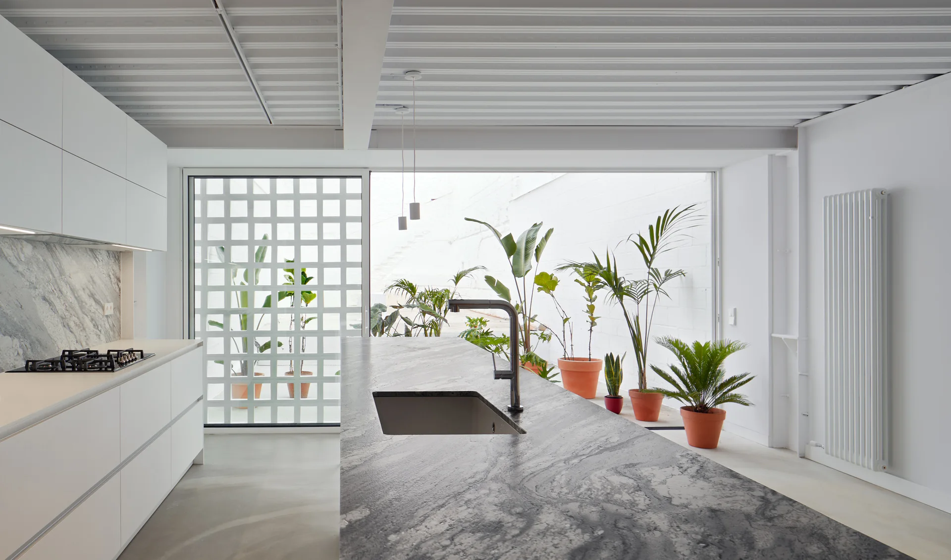 A minimalist kitchen with a veined grey stone island, white cabinetry, a corrugated metal ceiling, and sliding glass doors opening to potted courtyard plants.