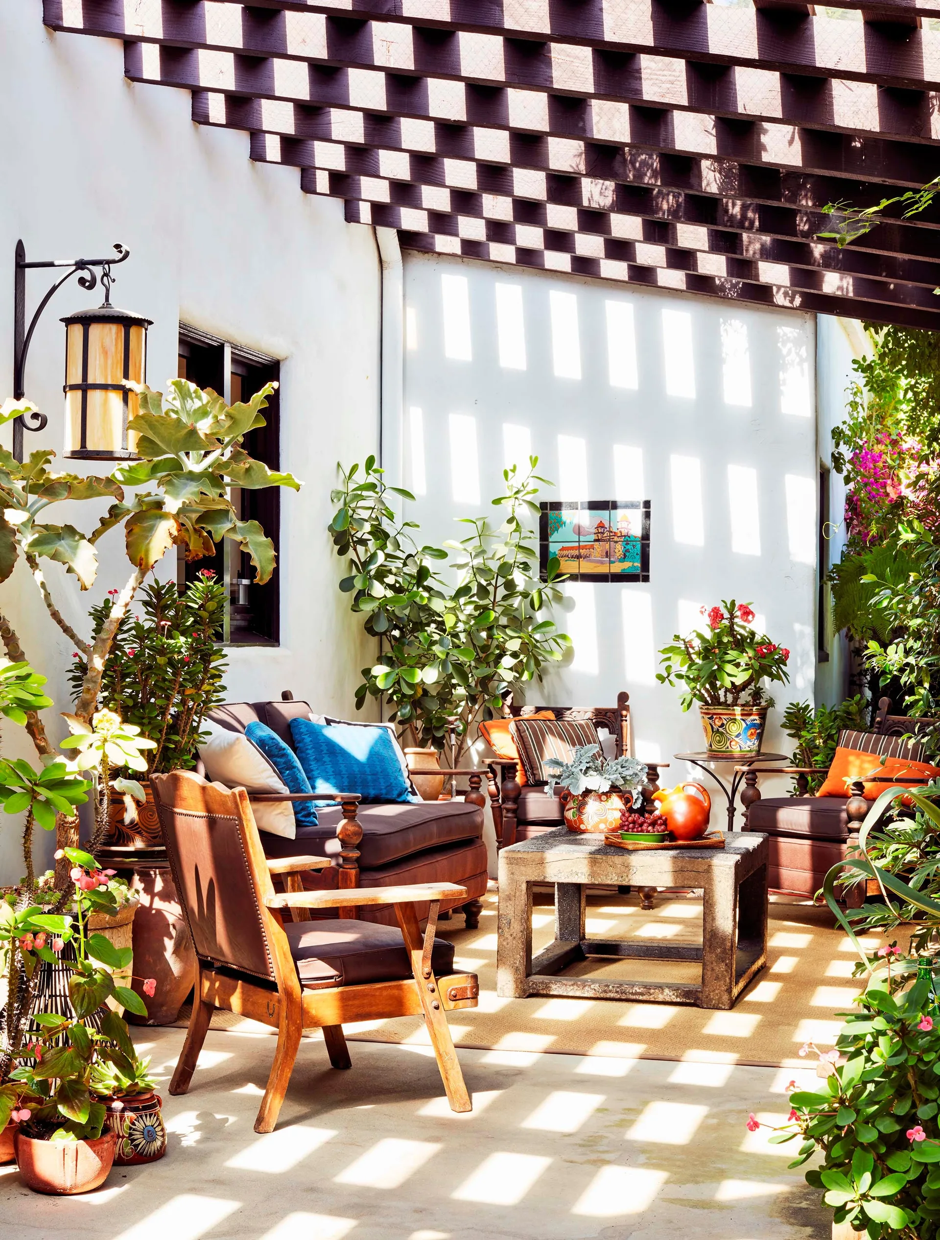 An outdoor patio features wood furniture with brown cushions under a dark wood pergola casting checkered shadows on a white wall.