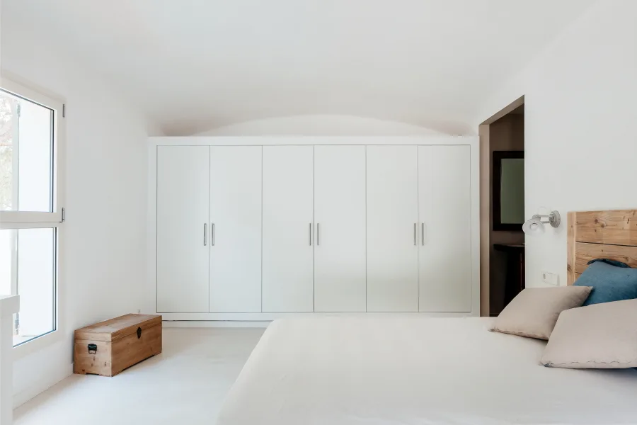 A white bedroom with a large built-in wardrobe, a bed with a wooden headboard, and a small wooden chest beneath a window.