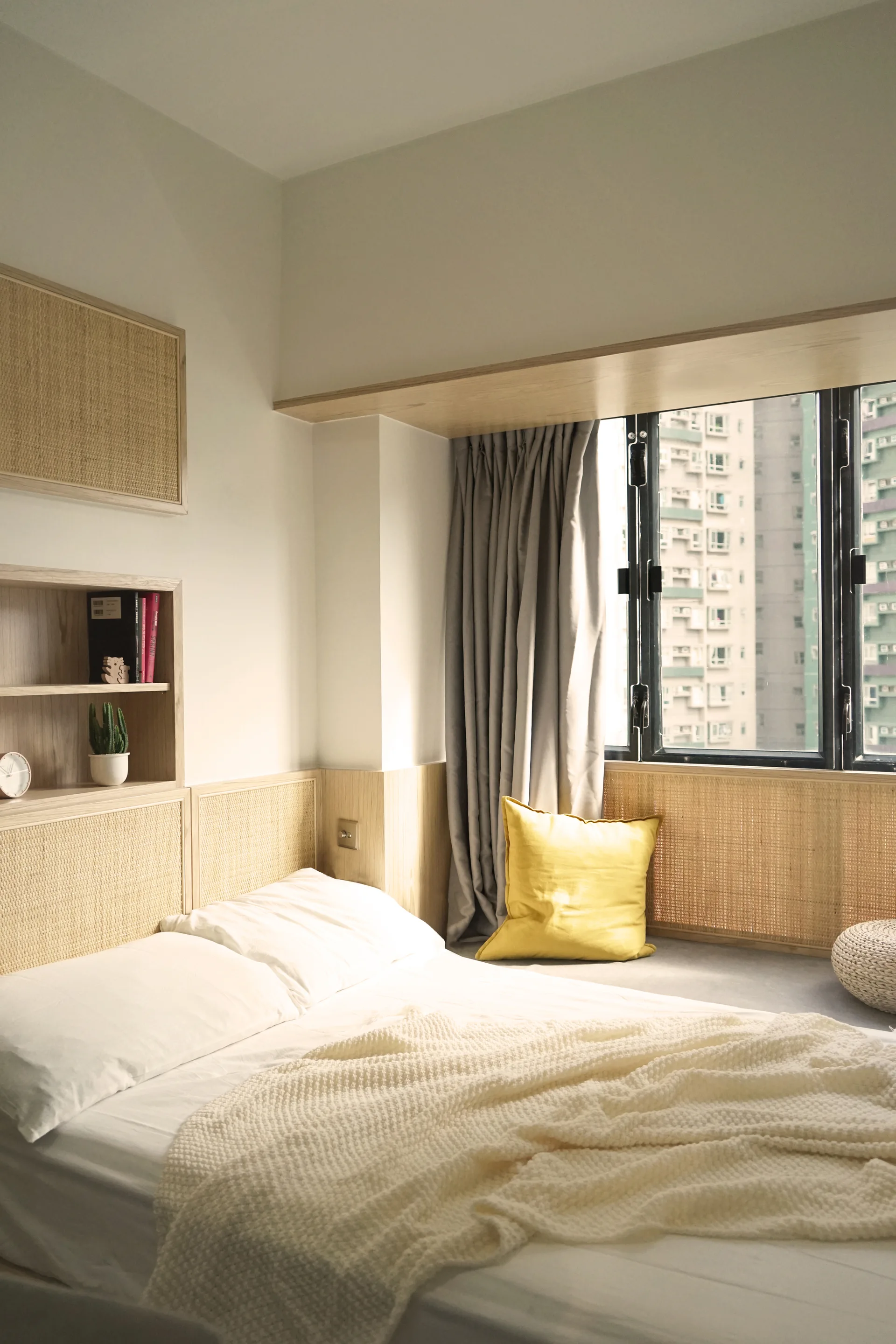 A low platform bed with white linens sits next to built-in light wood shelving and textured rattan wall paneling below a bright window.