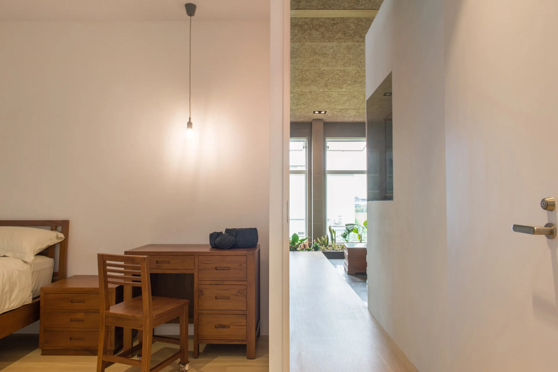 A minimalist bedroom featuring a wooden bed, nightstand, and desk with a matching chair, looking out towards a bright hallway.