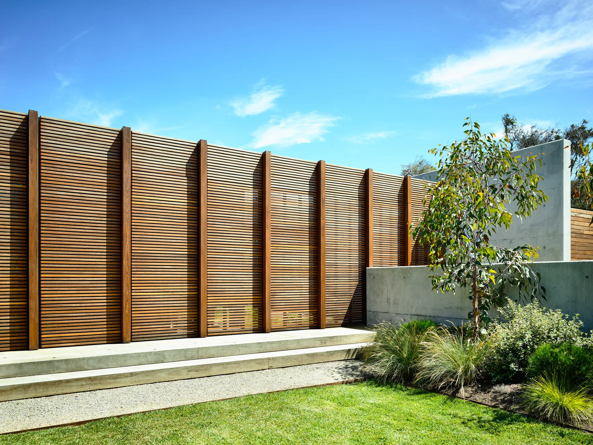 A modern outdoor garden featuring a tall horizontal slatted wood fence with vertical timber posts next to a grey concrete wall and green lawn.