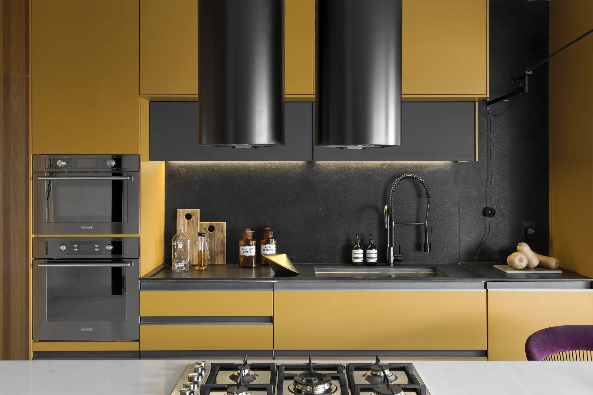 A modern kitchen features matte mustard yellow cabinets, a textured black backsplash, stacked wall ovens, and dual black cylindrical range hoods.