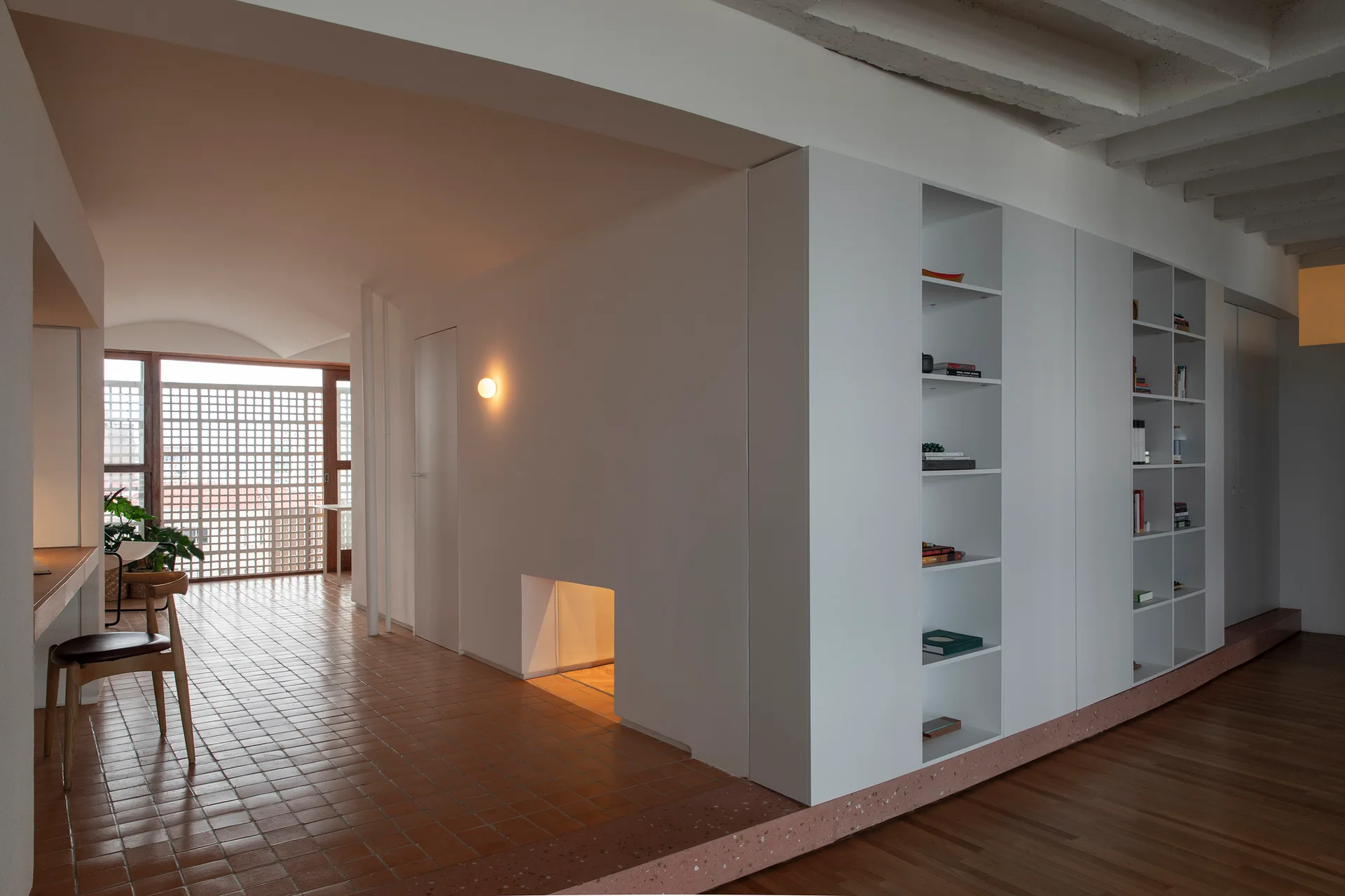 A contemporary interior hallway features white built-in storage shelves, terracotta tile floors, and a warm glowing wall sconce.