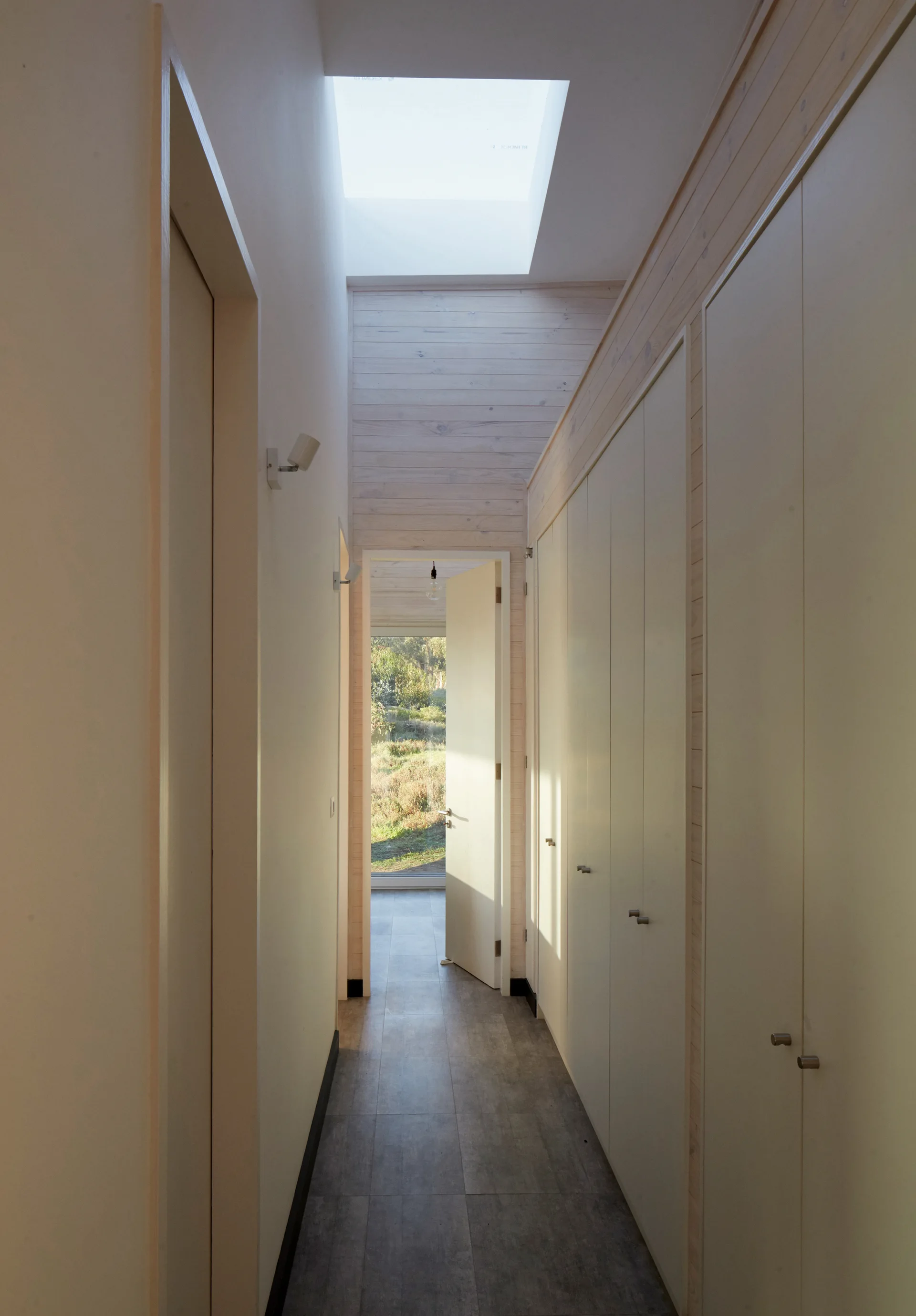 A narrow modern hallway with a large rectangular skylight, built-in floor-to-ceiling closets, and an open door showing grassy hills outside.