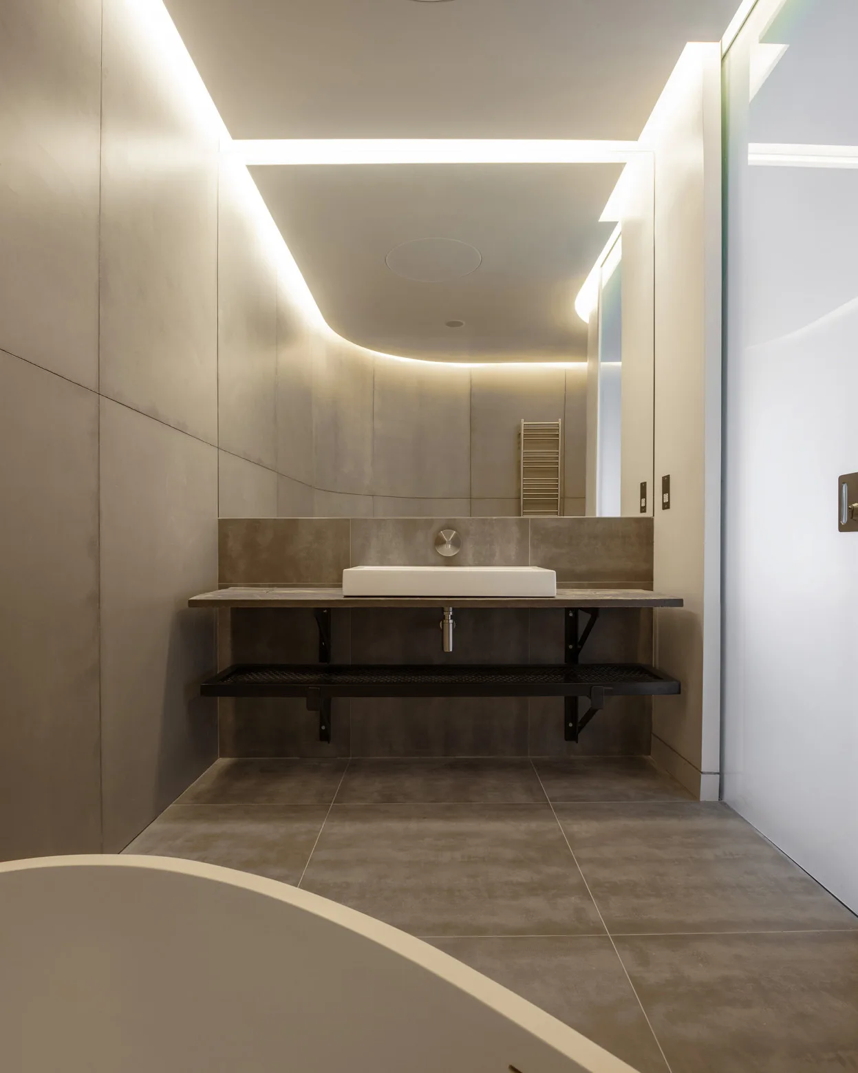 A minimalist bathroom featuring large-format gray tiles, a wall-mounted vanity with a white vessel sink, and glowing cove lighting along the ceiling.