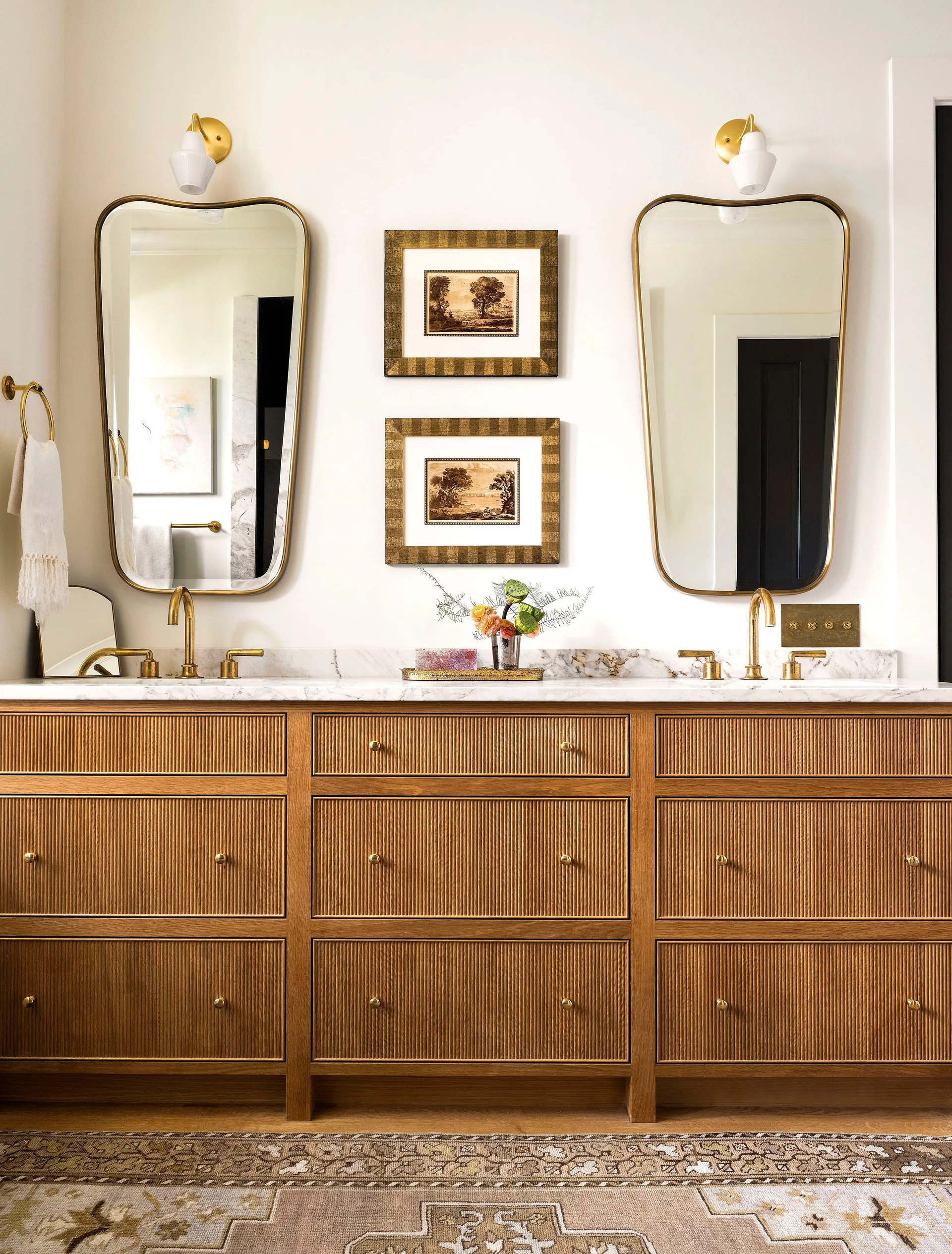 A double bathroom vanity features fluted wood drawers, a white marble countertop with two brass faucets, organic-shaped brass mirrors, and brass sconces.