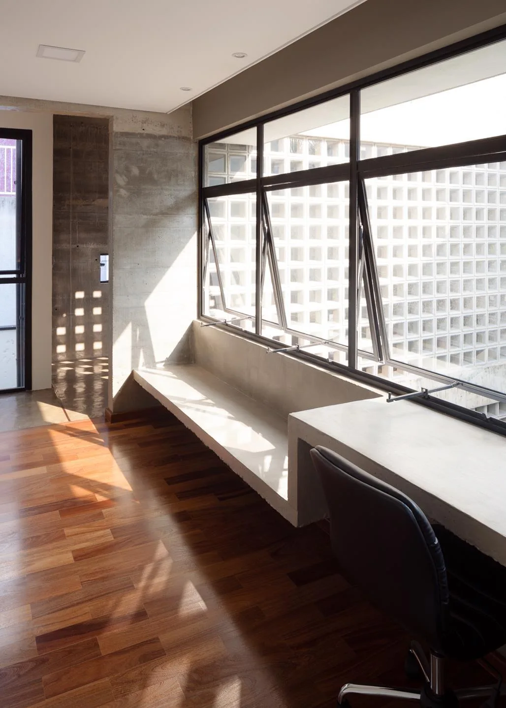 A minimalist home office featuring a built-in concrete desk beneath a large dark-framed window, set against warm wood floors and exposed concrete walls.