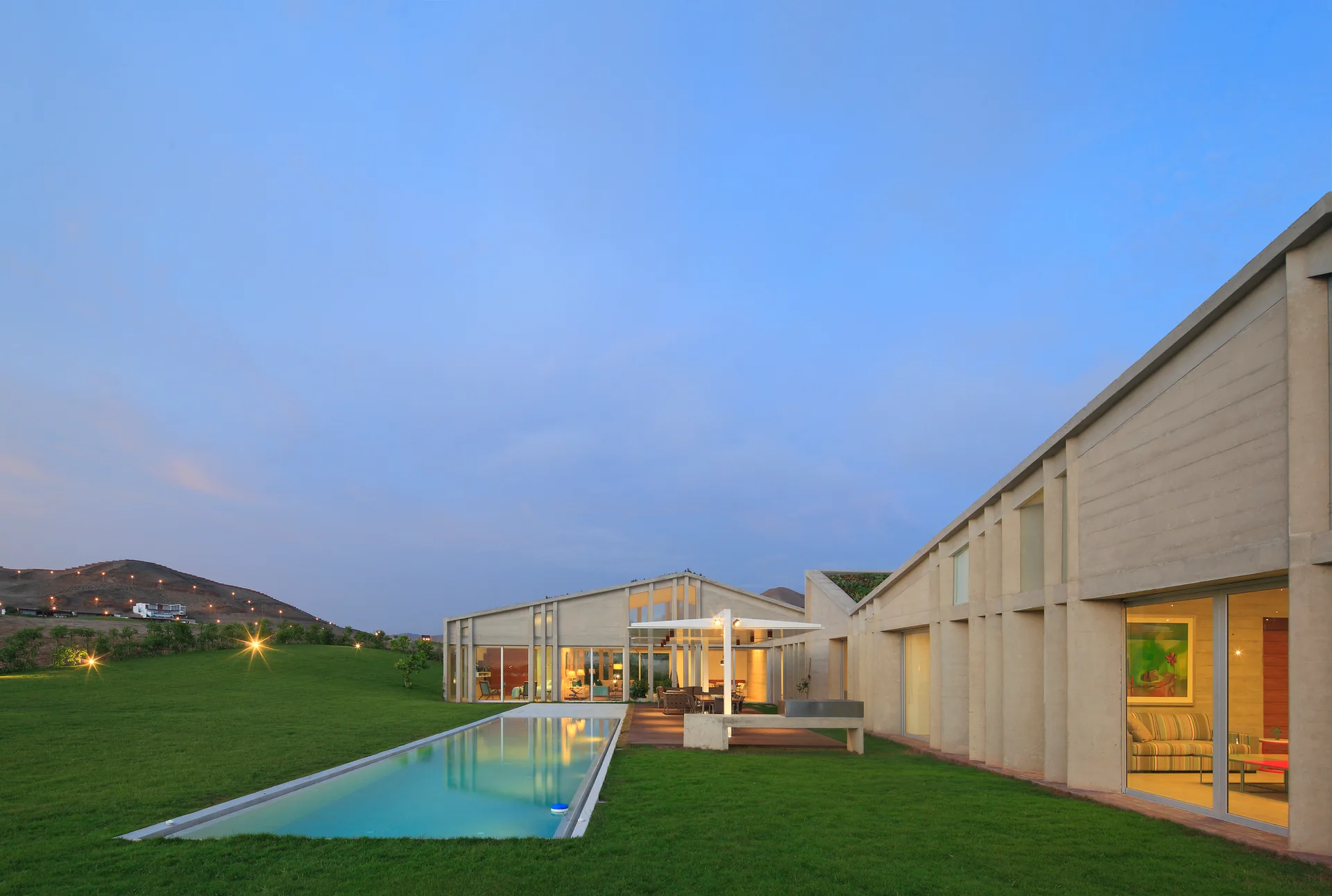 A modern concrete house features large glass doors, an outdoor wooden deck, and a long rectangular pool set in a large green lawn at dusk.