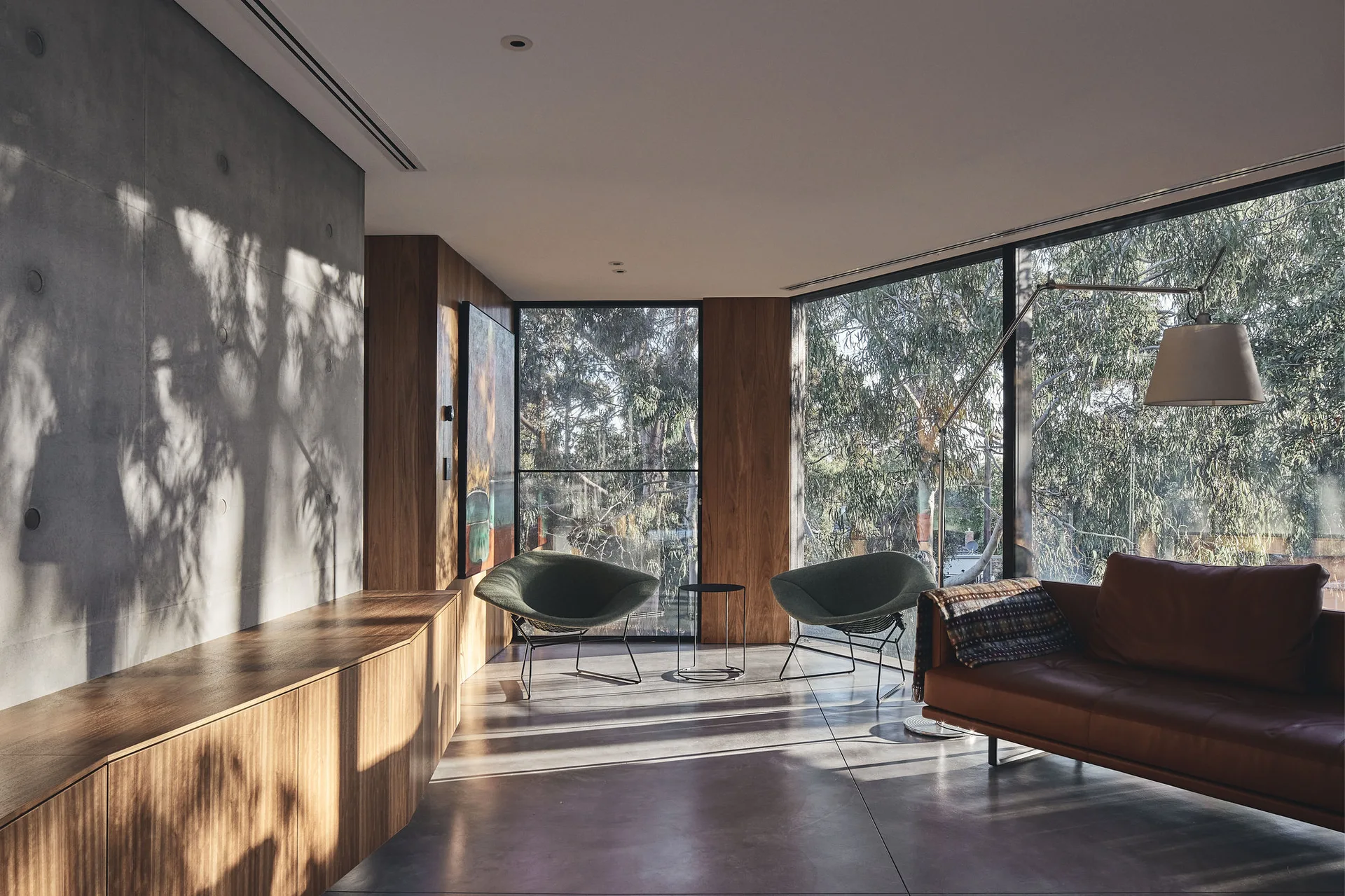 A minimalist living room features a brown leather sofa, green armchairs, an exposed concrete wall, and large windows looking out at trees.