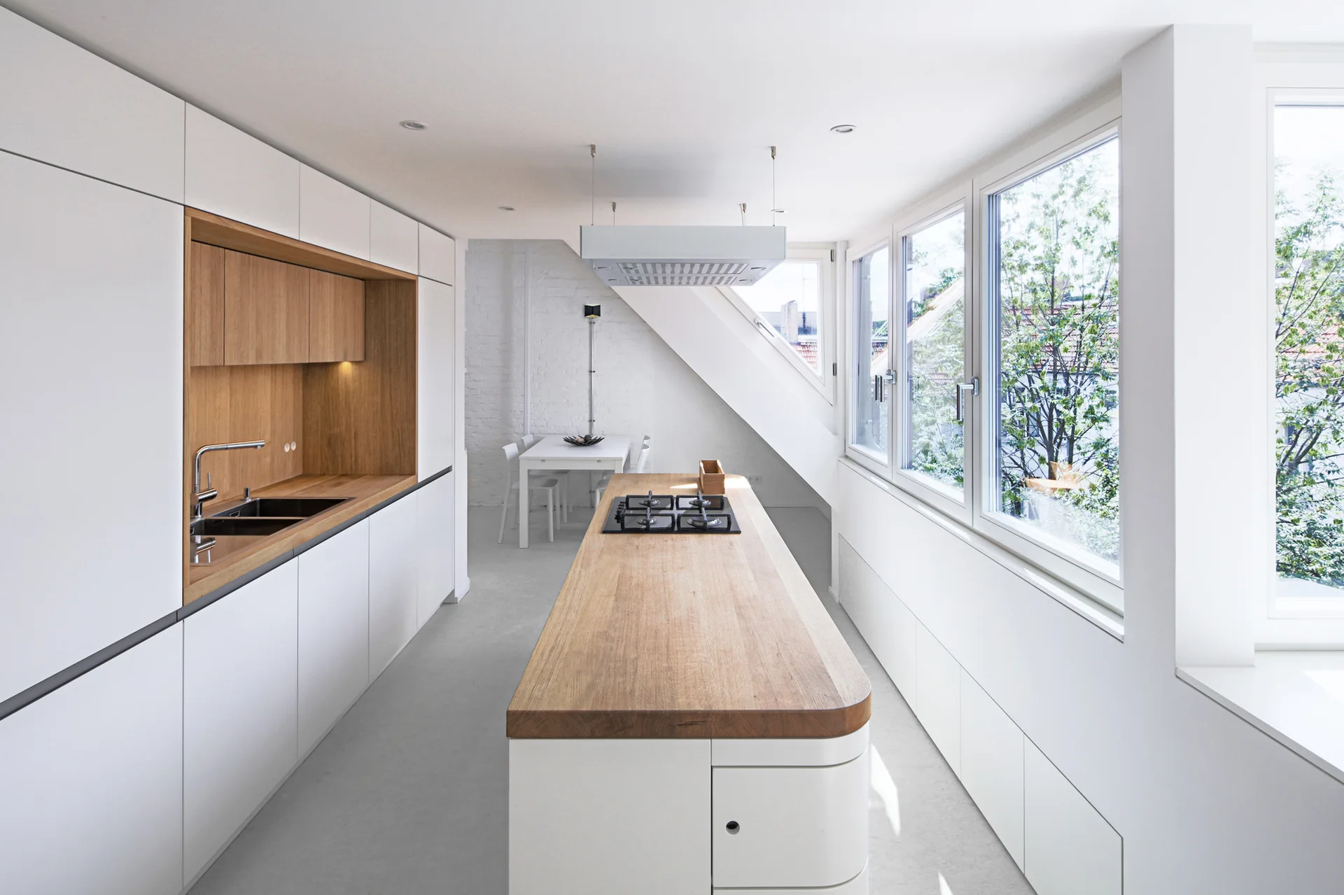 A minimalist attic kitchen features integrated white cabinets, a long kitchen island with a natural wood countertop, and a row of large side windows.