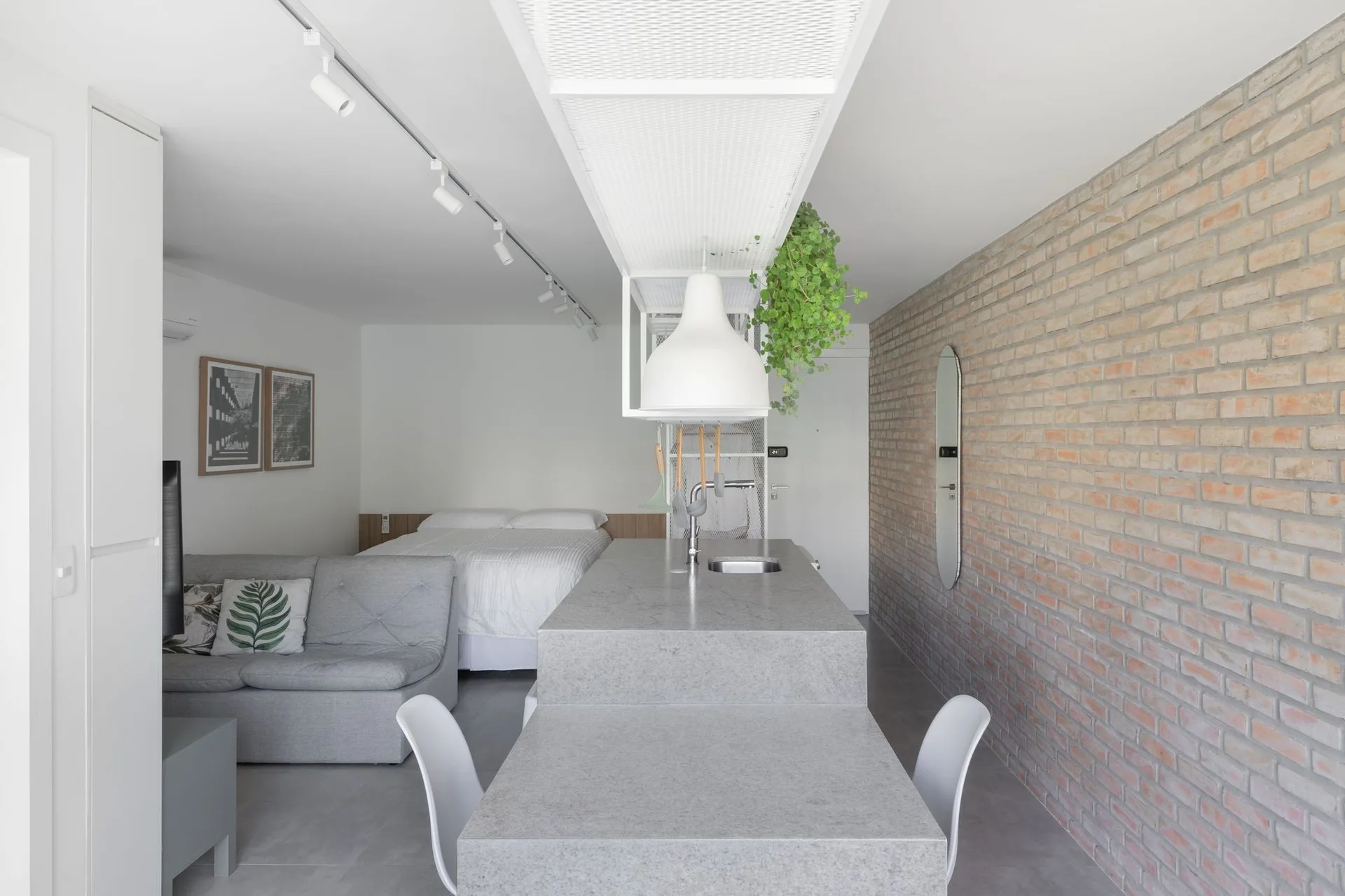 A minimalist studio apartment featuring a grey stone kitchen island, an exposed brick accent wall, and a grey sofa.