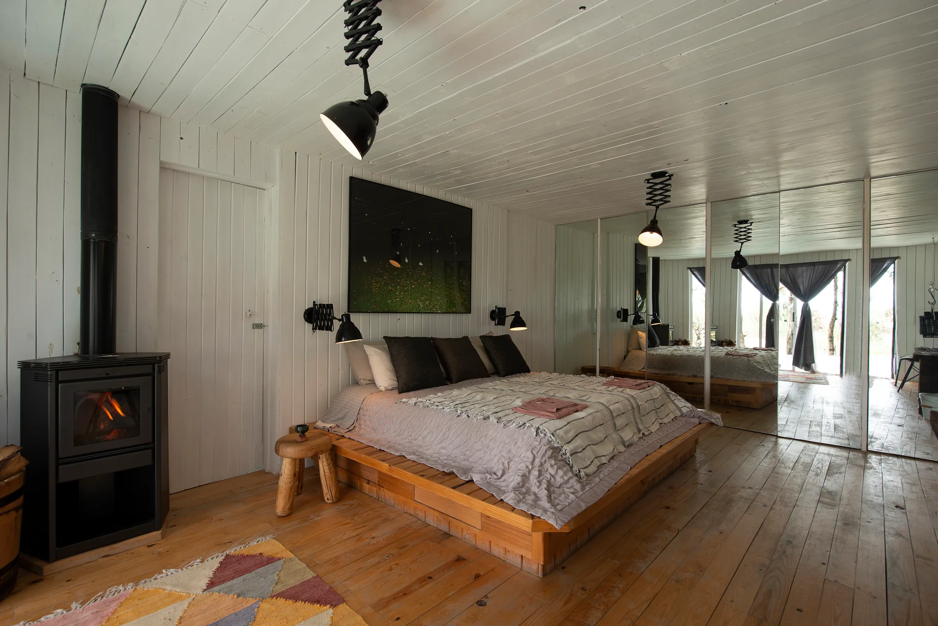 A rustic bedroom features a wooden platform bed next to a black wood stove with white vertical plank walls and a mirrored wardrobe.