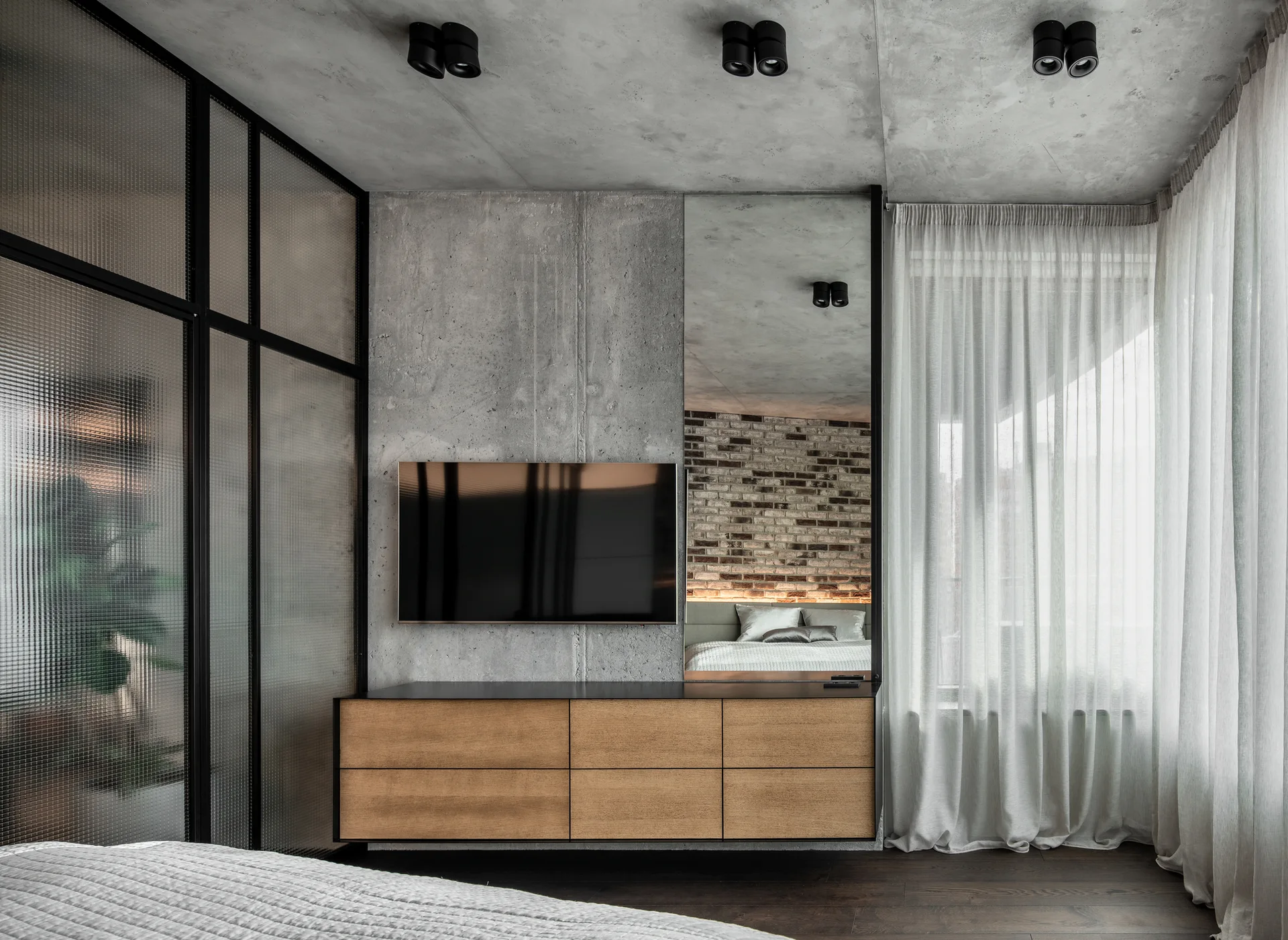A contemporary bedroom features an exposed concrete wall with a mounted TV, a floating wood dresser, and a textured glass partition.