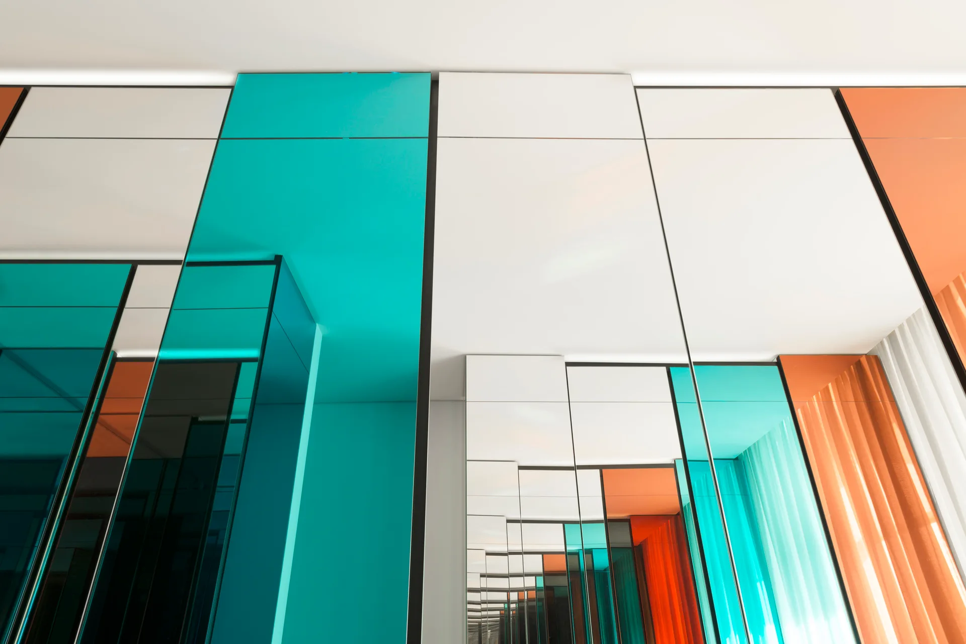 Architectural detail of a wall clad in glossy, geometric glass panels in teal, orange, and white under a plain ceiling.