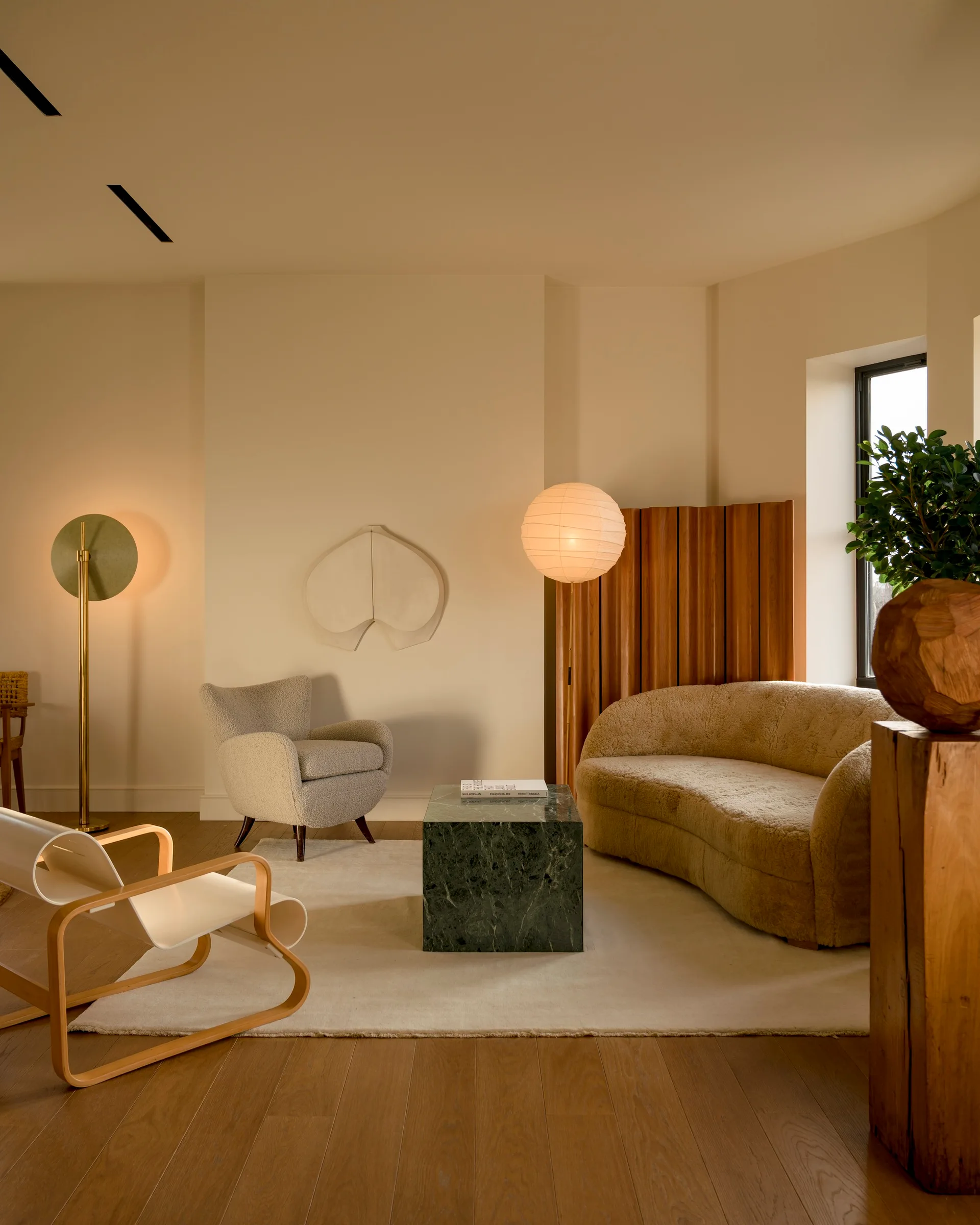 An organic modern living room featuring a curved boucle sofa, a dark green marble cube coffee table, a spherical paper floor lamp, and a wooden divider.