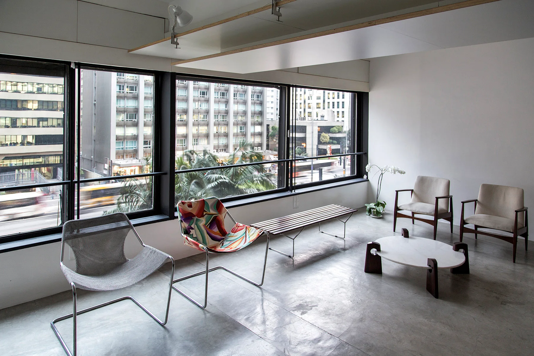 A minimalist living room features a grey concrete floor, diverse modern chairs, an oval coffee table, and large windows looking out on city buildings.