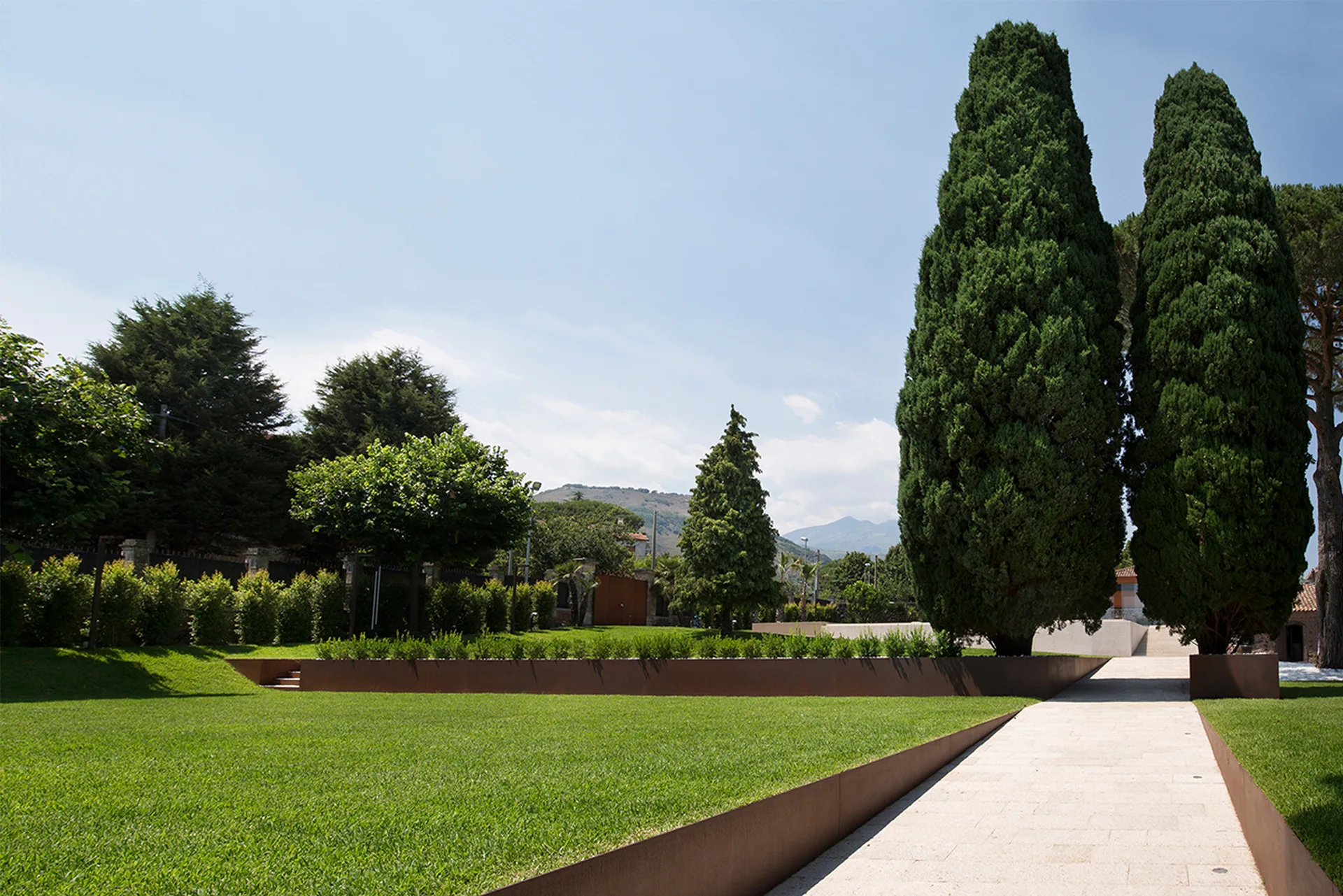 A paved stone pathway cuts through a manicured green lawn flanked by tall cypress trees and weathered rust-colored metal retaining borders.