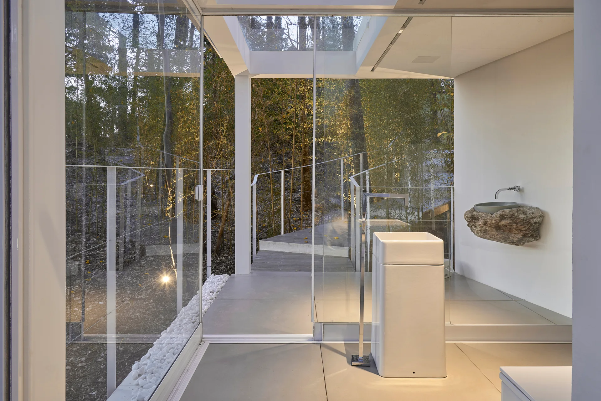 A minimalist bathroom with floor-to-ceiling glass walls, featuring a white pedestal sink and a rustic stone basin mounted on a plain white wall.