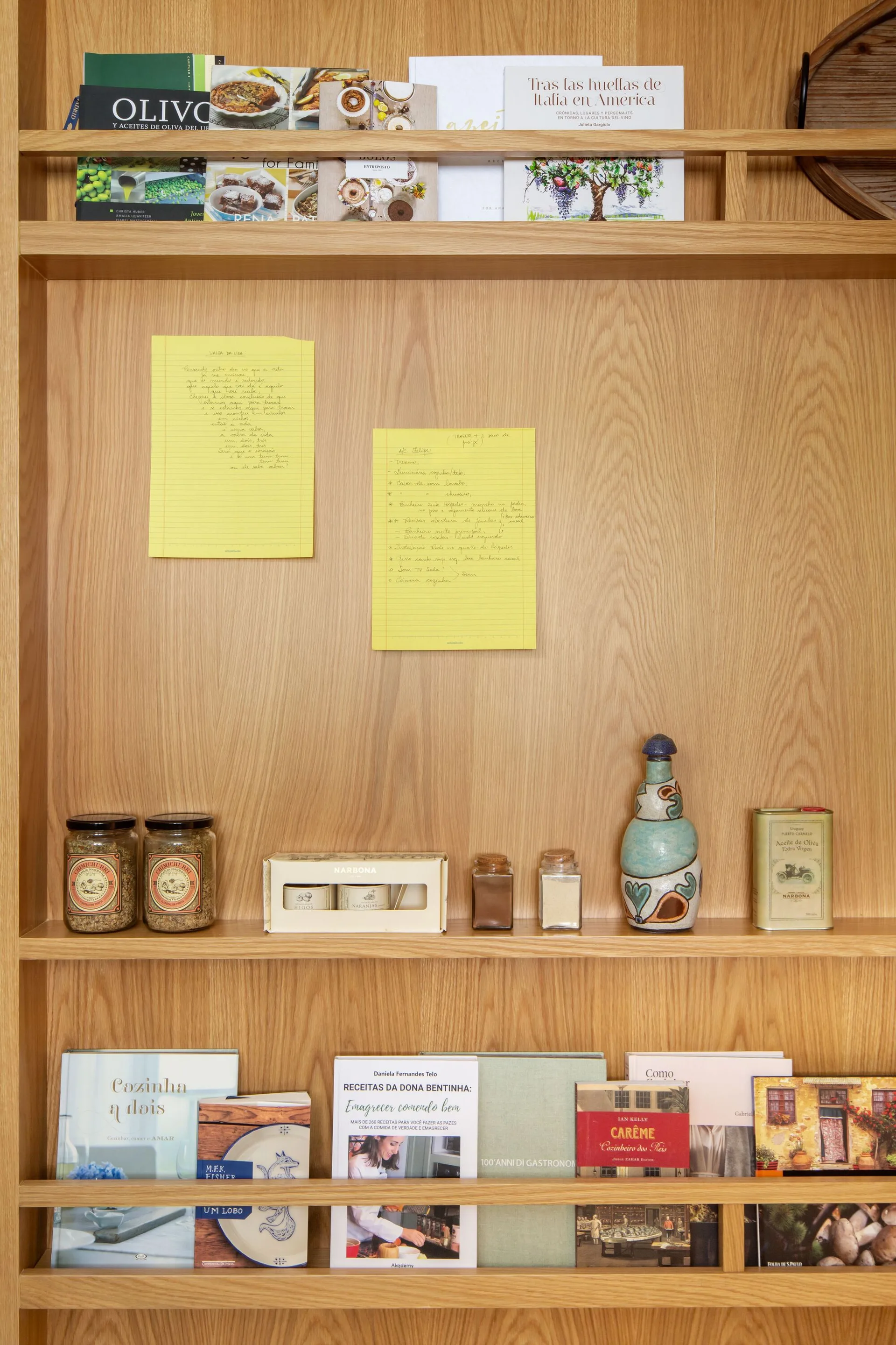 A light oak wooden shelving unit features front-facing cookbooks behind thin wooden guard rails, alongside glass spice jars and handwritten yellow notes.