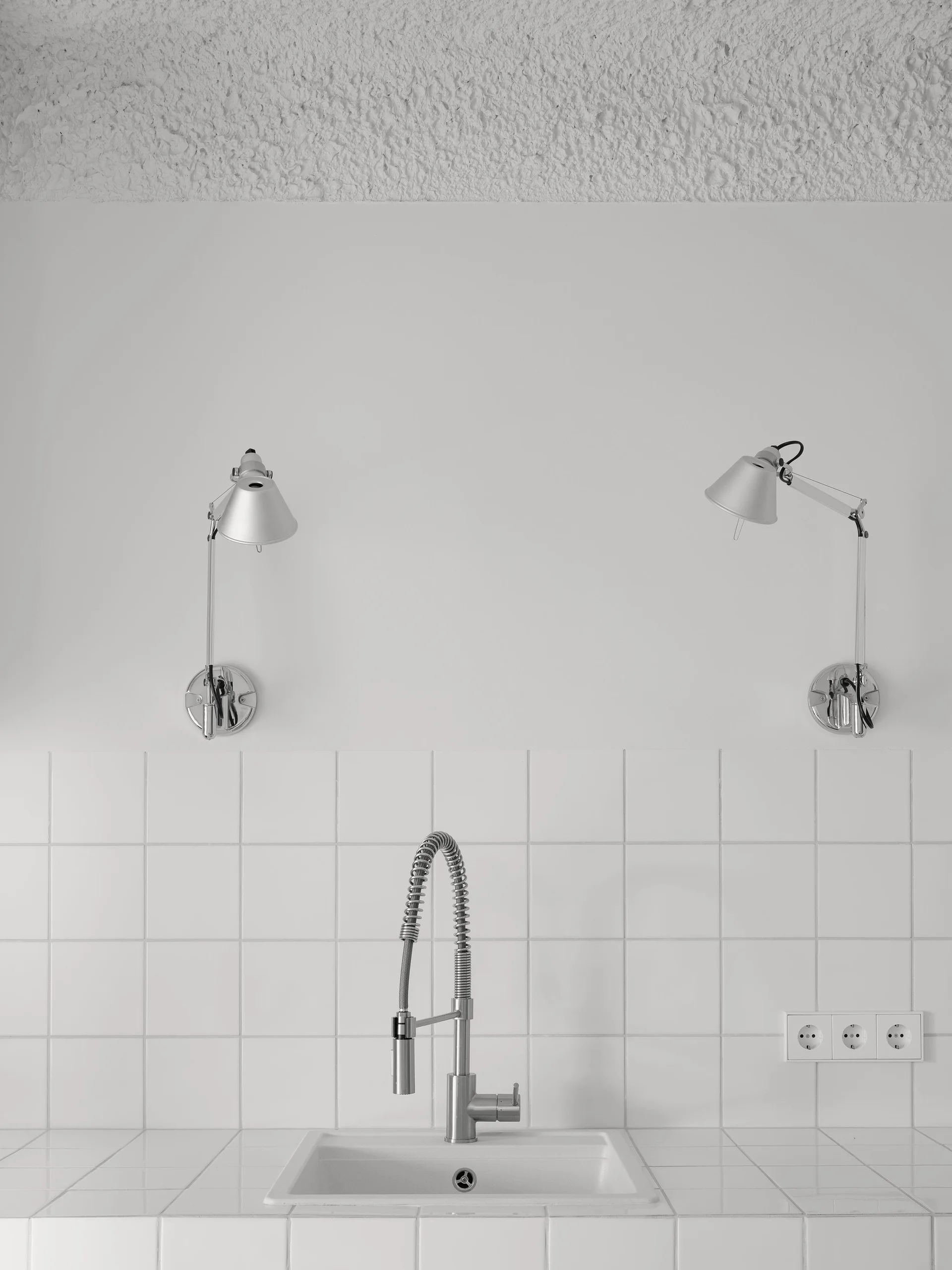 A chrome pull-down faucet and integrated white sink are surrounded by white square wall tiles beneath two adjustable silver wall-mounted lamps.