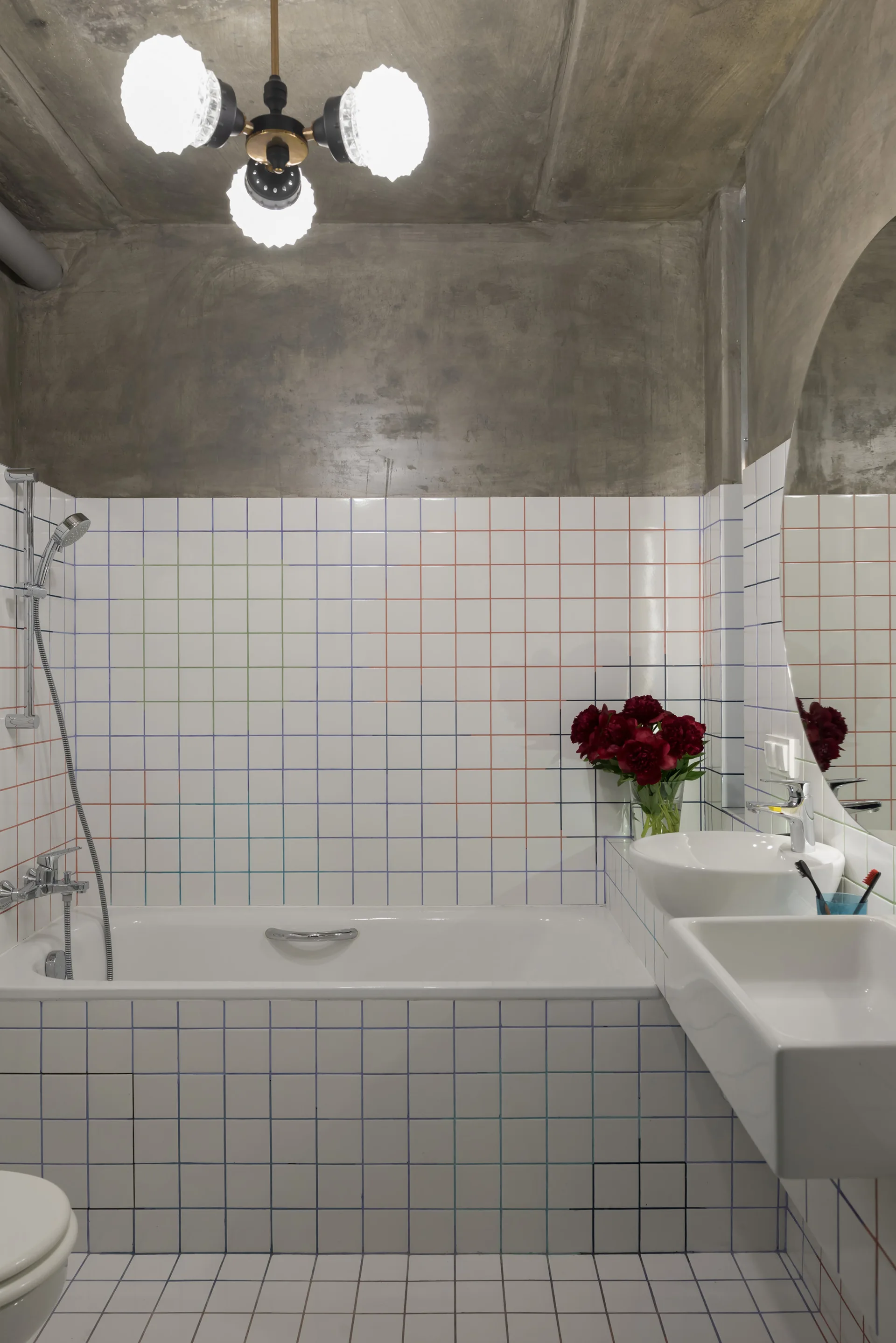 A minimalist bathroom featuring a white tiled bathtub and walls with multi-colored grout, a concrete ceiling, and a modern light fixture.