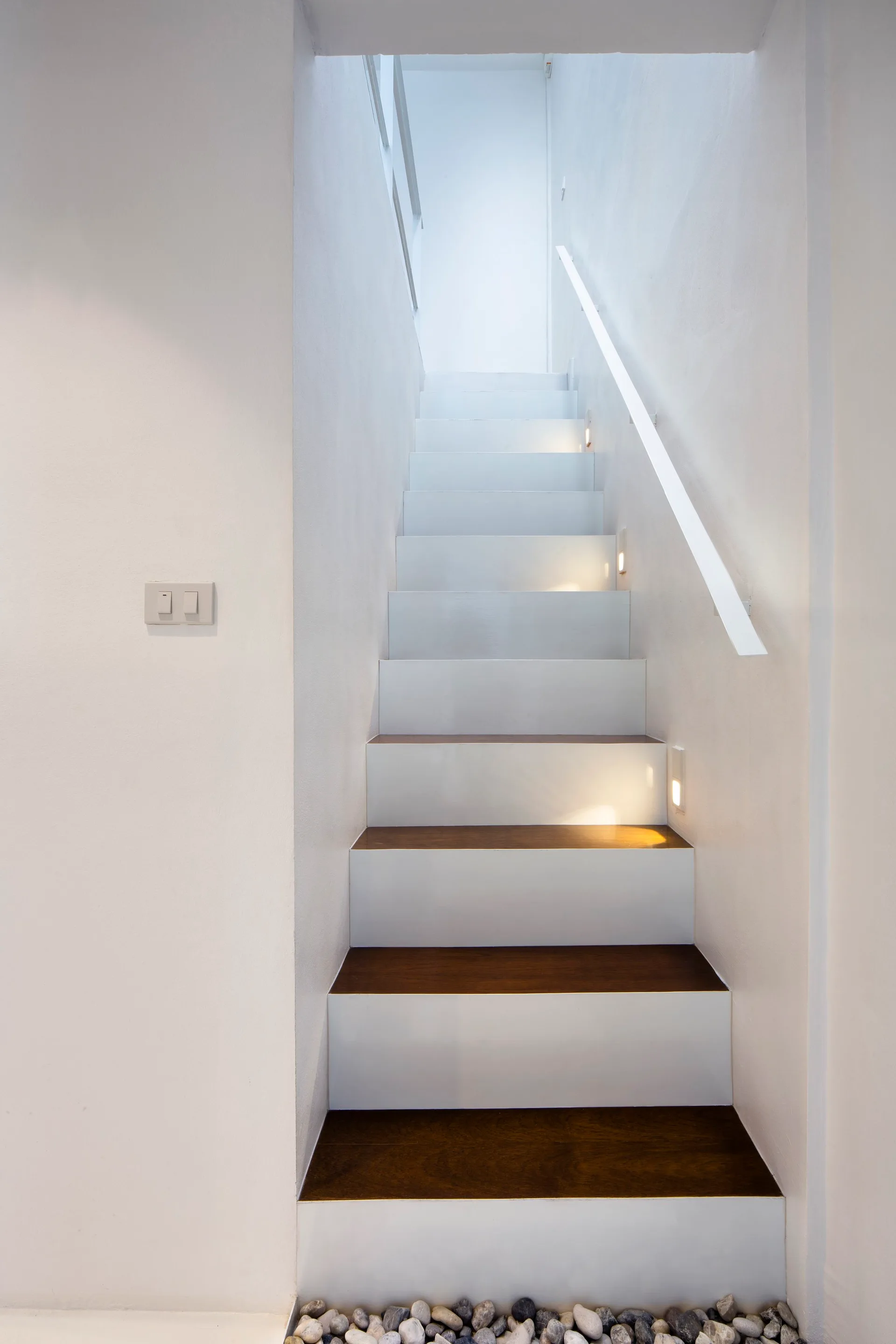 A minimalist white staircase features dark wood treads, integrated wall-mounted step lights, and a small bed of pebbles at the base.