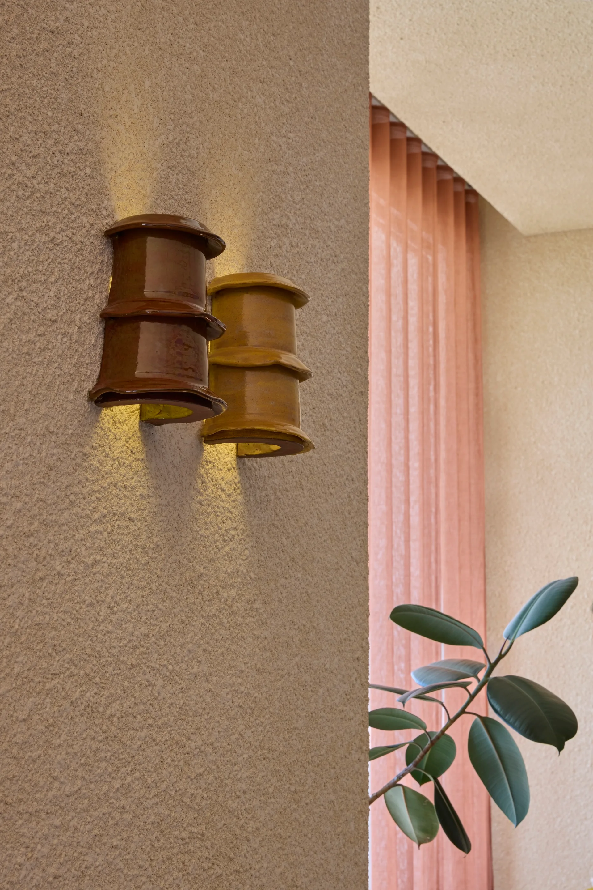Two stacked ceramic wall sconces in brown and ochre glaze cast a warm glow on a rough stucco wall next to a pink curtain.