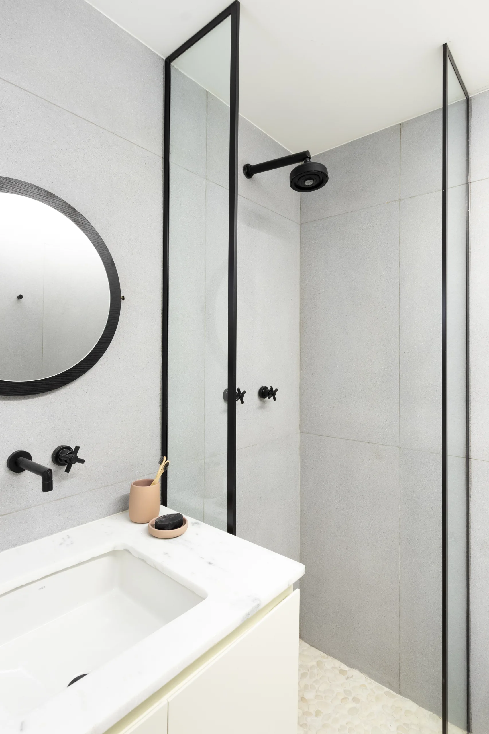 A modern bathroom features grey tiled walls, a white marble vanity with a black faucet, and a glass-enclosed shower with a black metal frame.