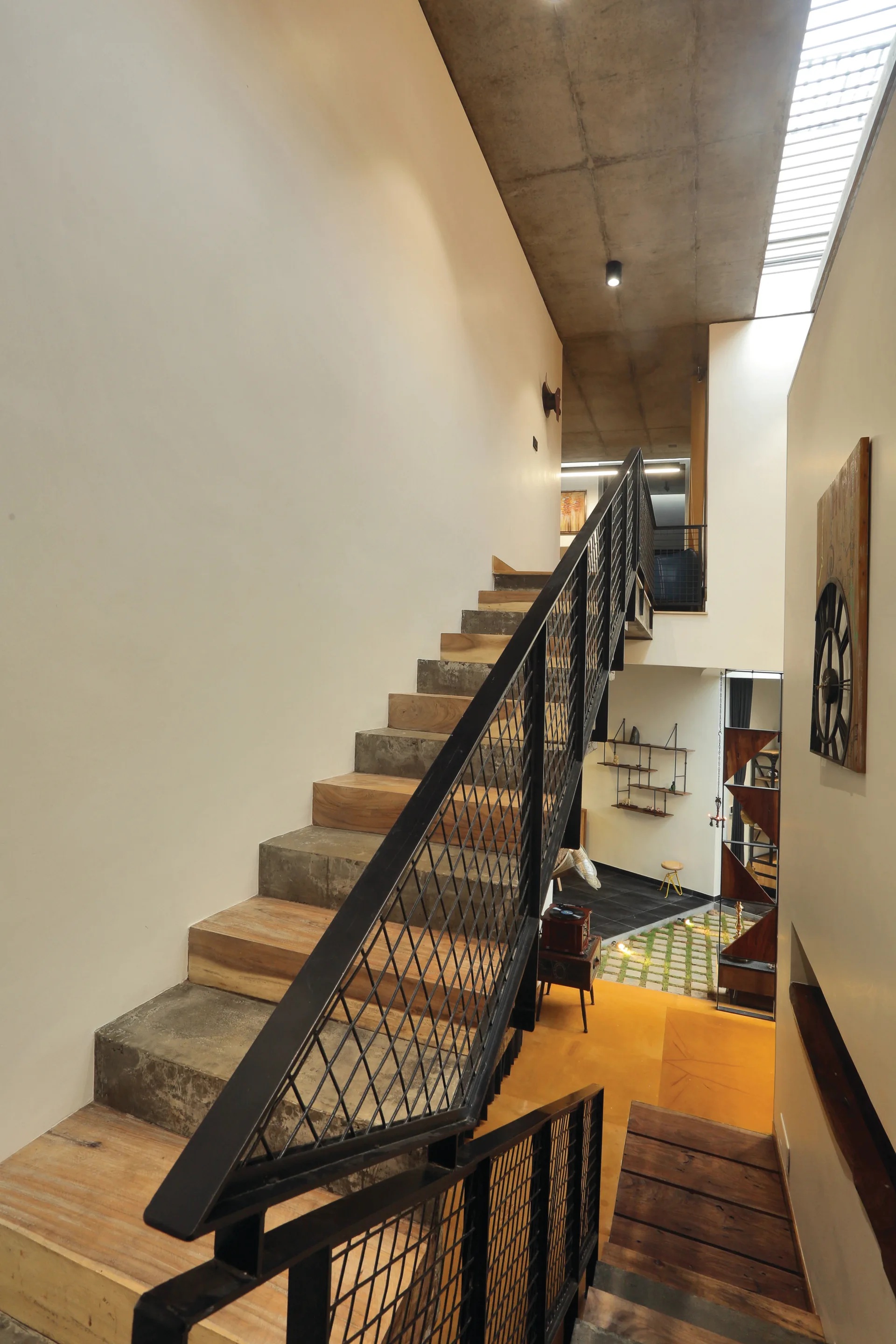 A multi-level staircase with alternating wood and concrete steps features a black metal mesh railing under a raw concrete ceiling and skylight.