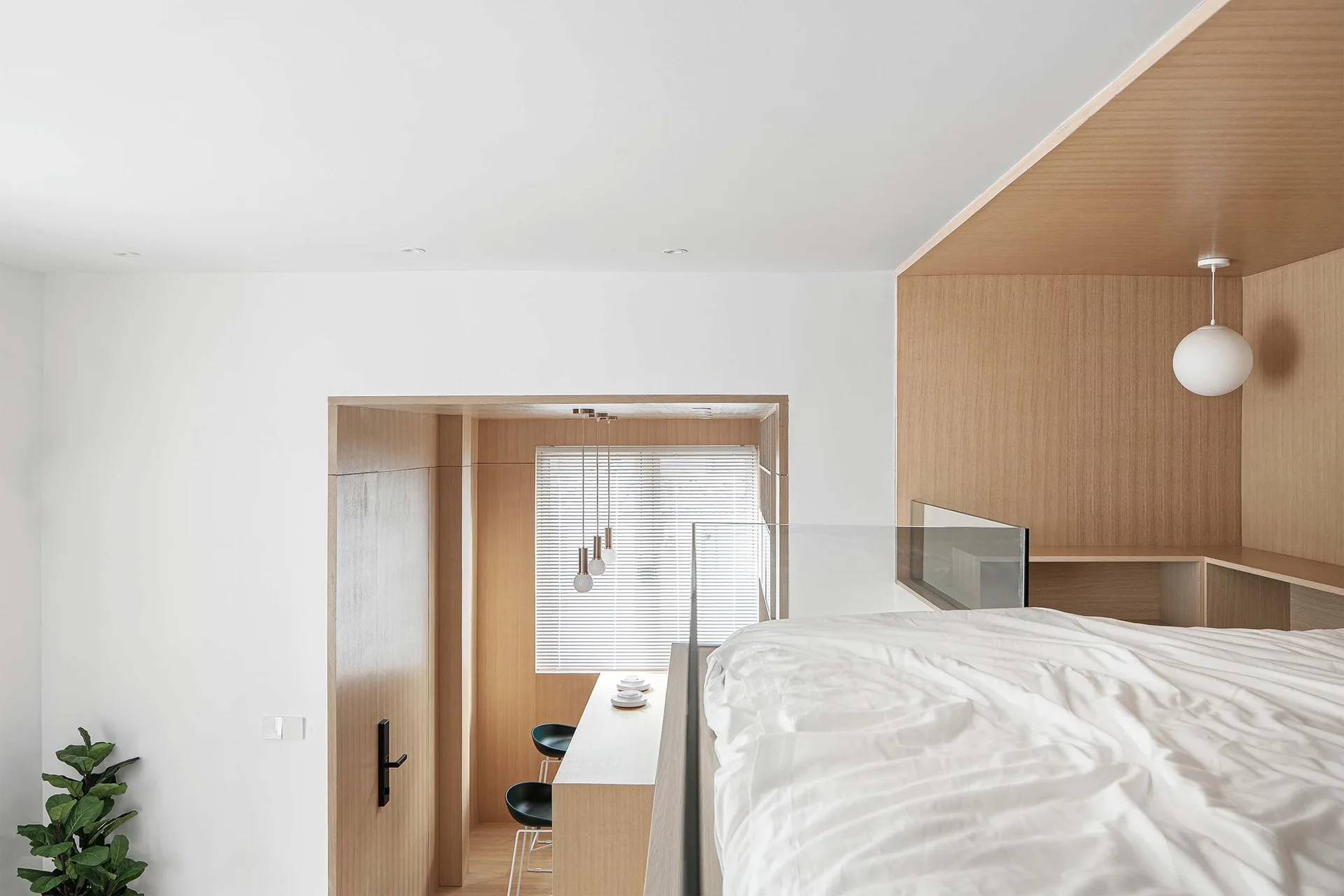 An elevated minimalist loft bed with white bedding features a clear glass railing and warm wood paneling next to a lower bar area with stools.