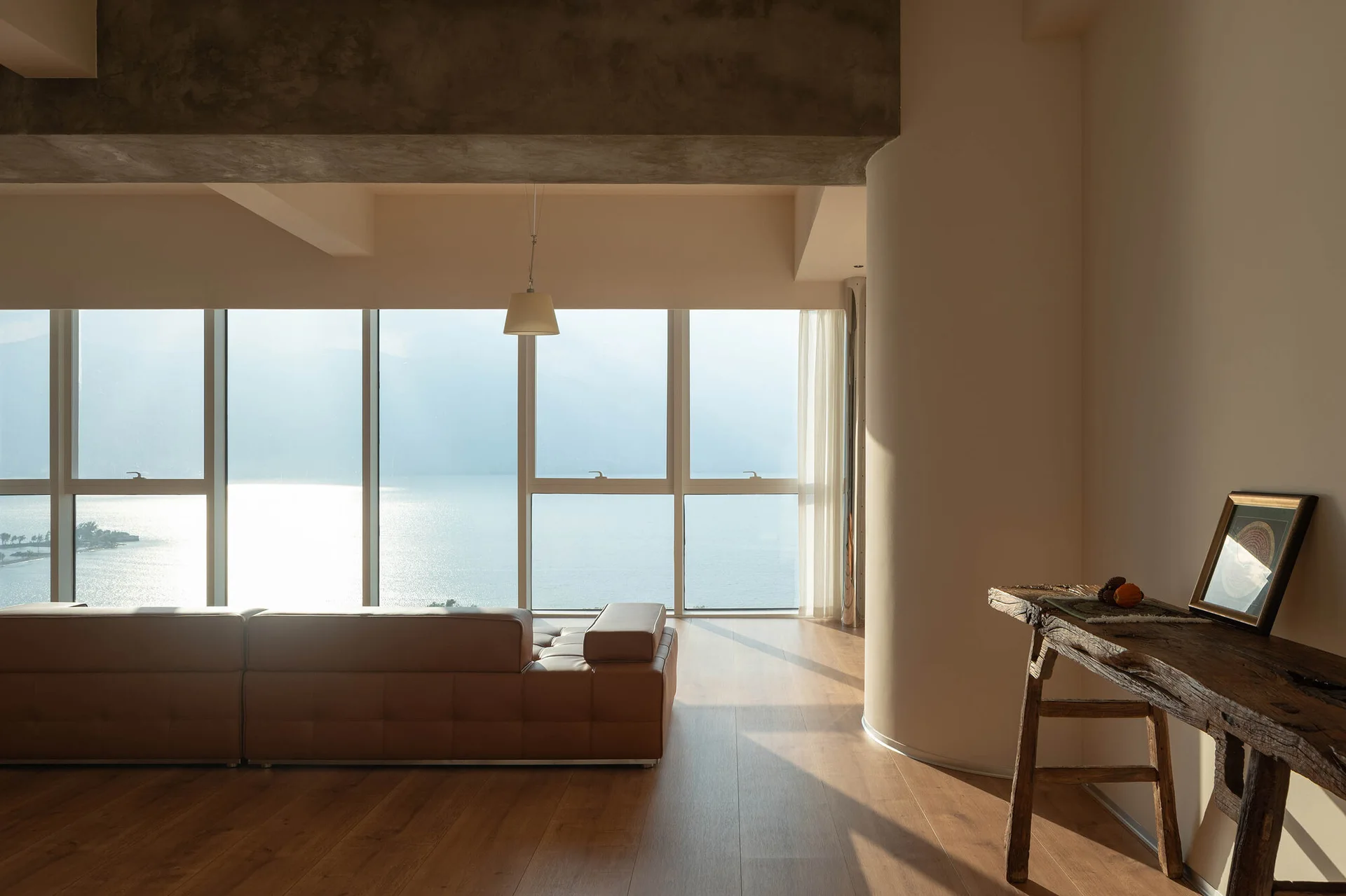 A minimalist living room features a brown leather sofa, a rustic wooden console table, and large windows looking out onto a bright body of water.