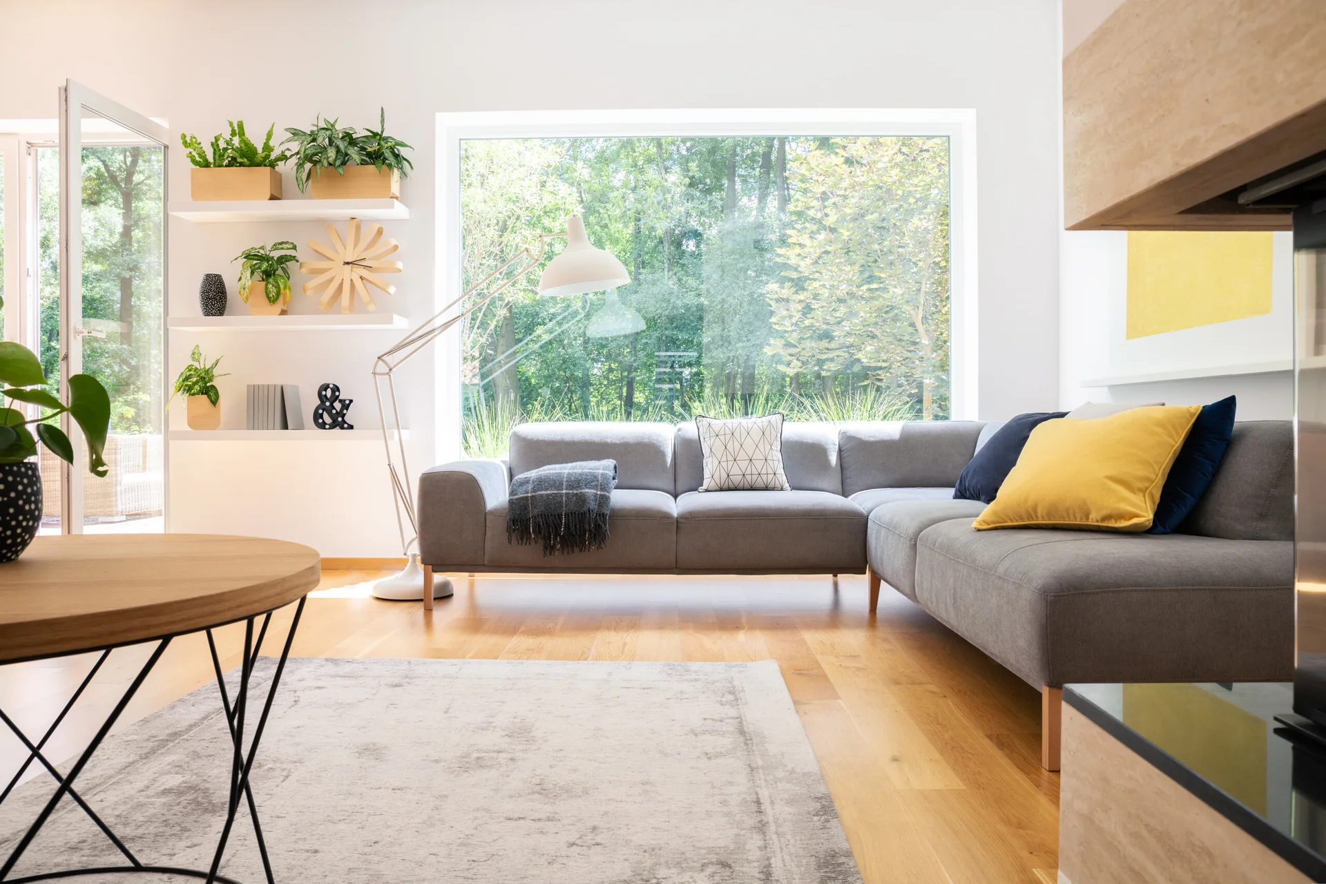 A bright living room features a grey sectional sofa, a round wooden coffee table, white floating shelves with plants, and a large picture window.