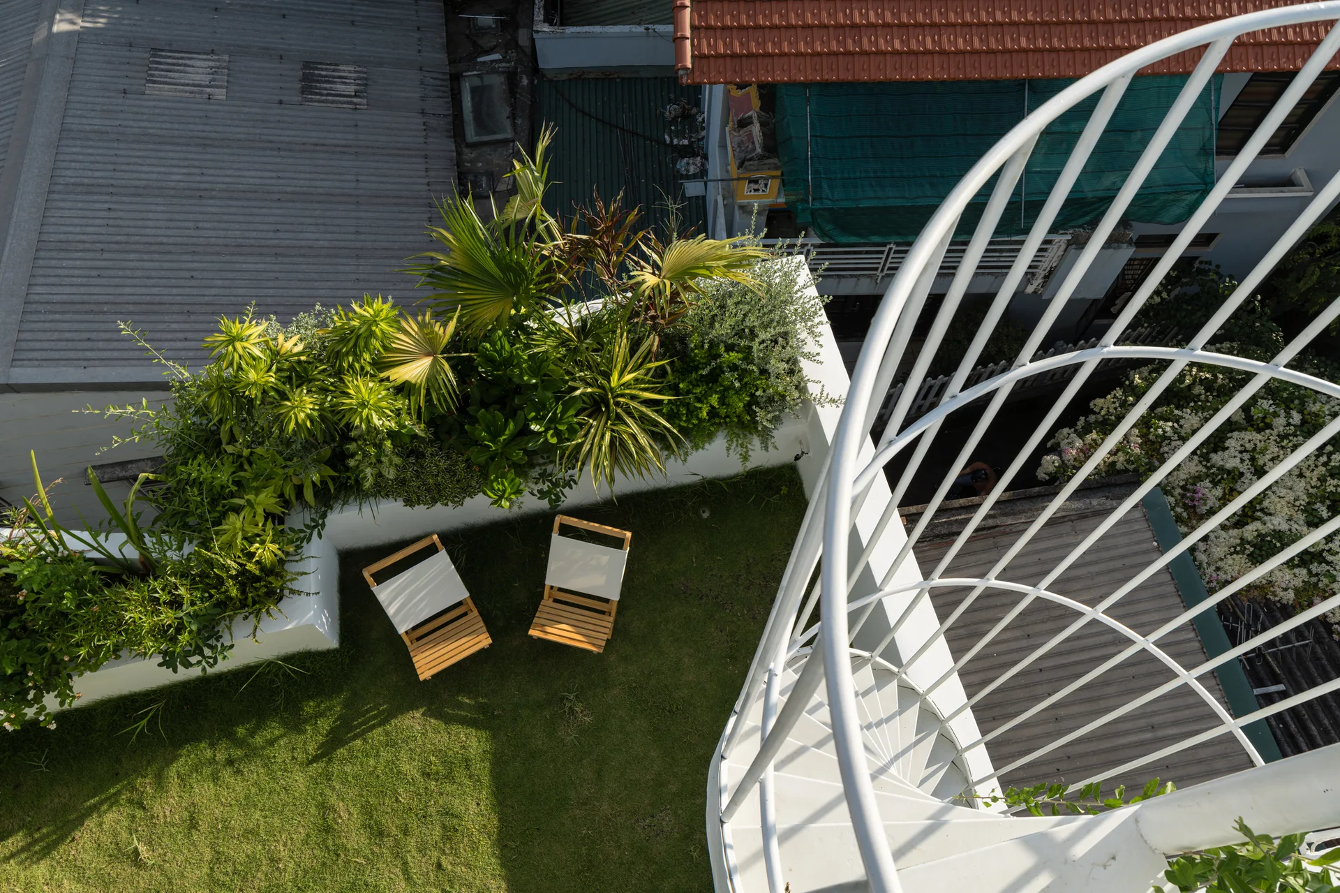 An overhead view of a rooftop lawn featuring two wood-and-canvas folding chairs next to a white metal spiral staircase and raised green planters.
