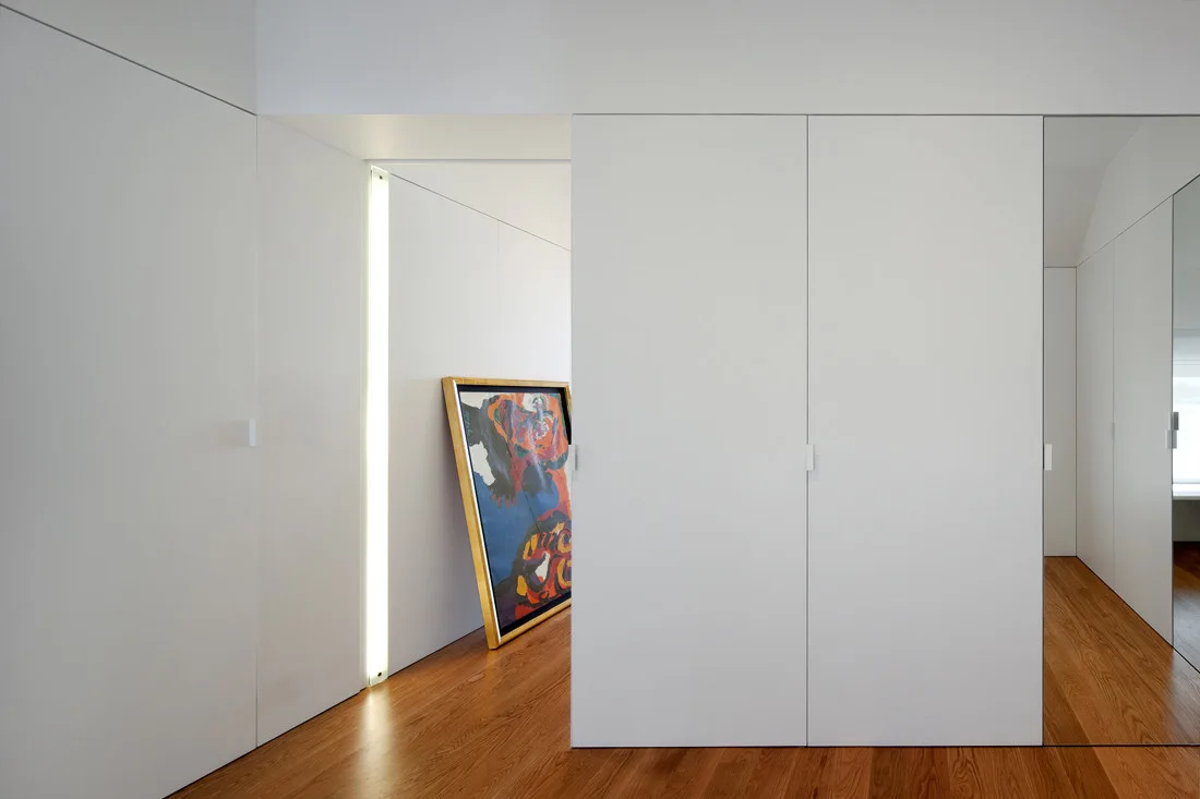 Minimalist hallway featuring seamless floor-to-ceiling white panels, warm wood floors, a vertical recessed light strip, and leaning colorful artwork.