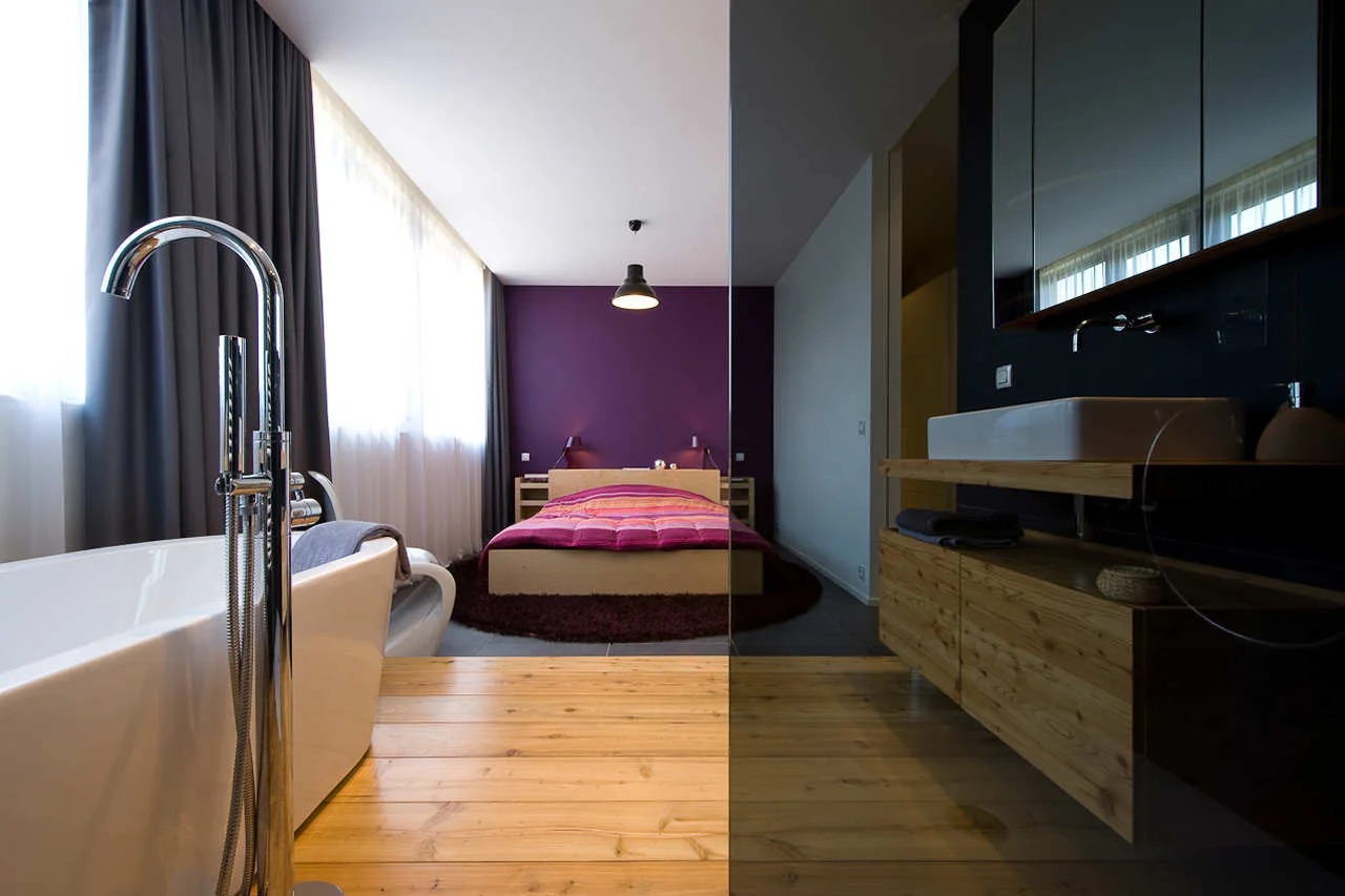 An open-concept master bedroom featuring a freestanding bathtub and wood vanity separated from the bed by a clear glass partition.