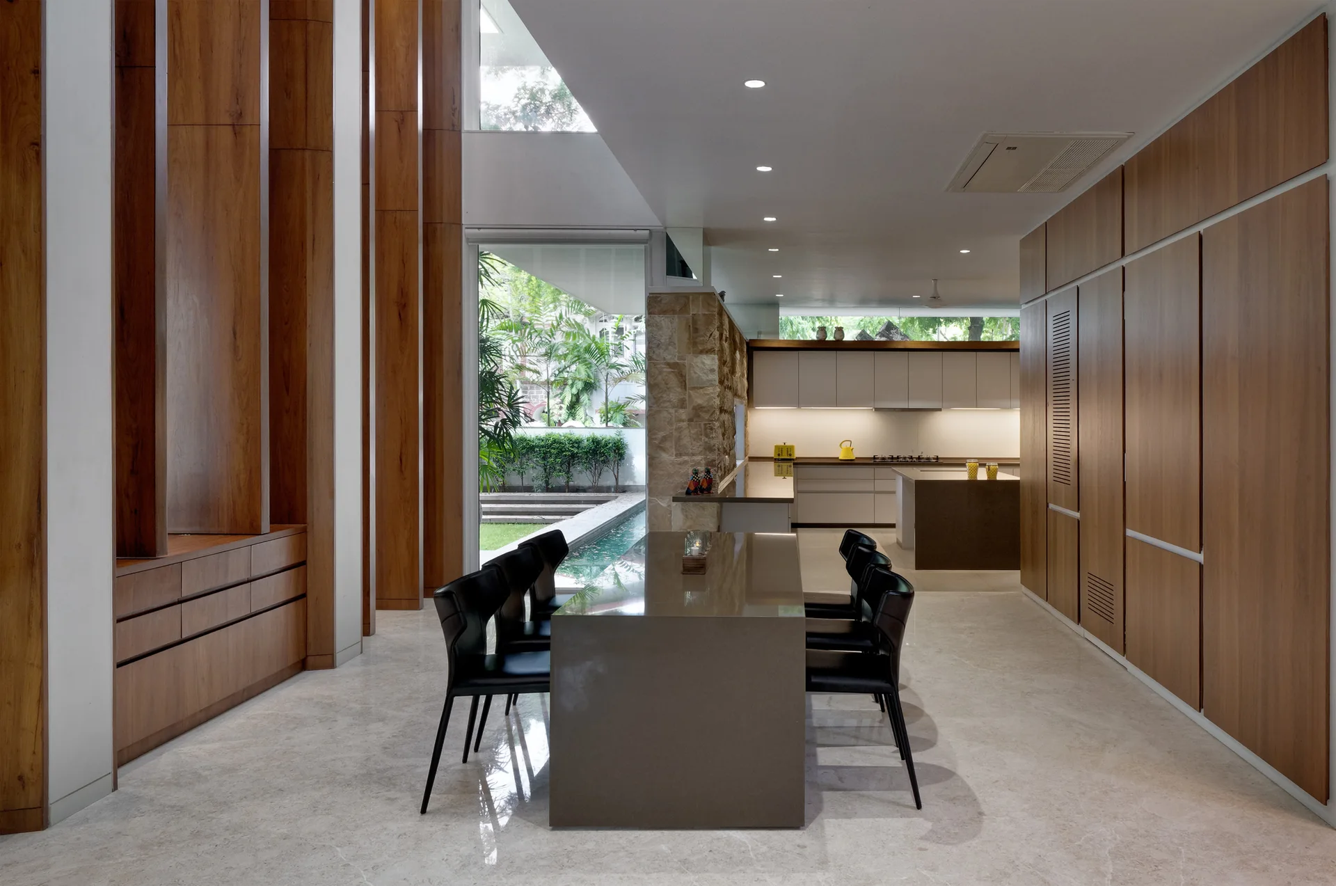 A modern open-plan dining room features a long grey table with black chairs, flanked by tall wood paneling and a kitchen in the background.