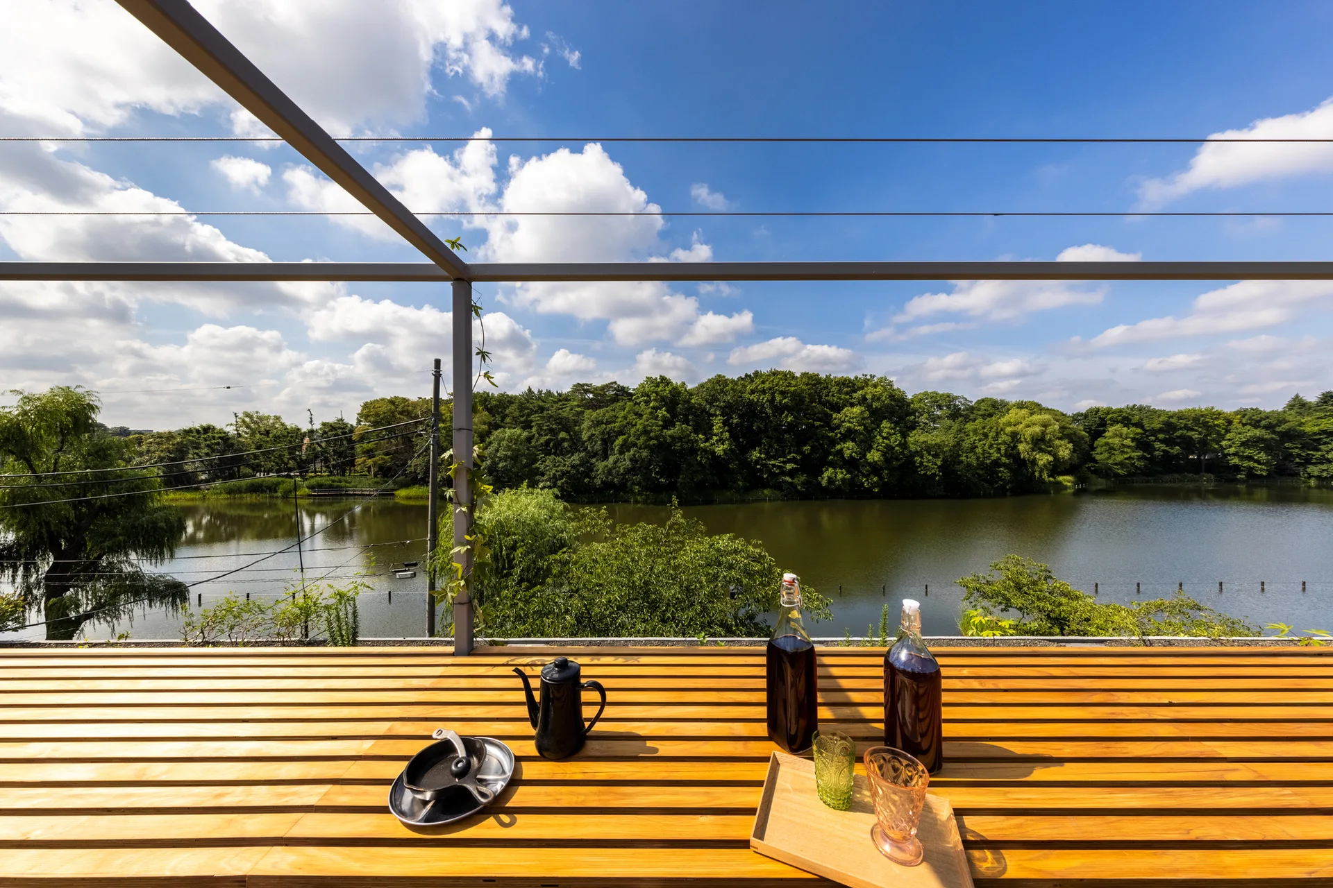 A wooden slatted terrace table set with a black kettle, bottles, and glasses overlooking a scenic lake and green forest under a blue sky.