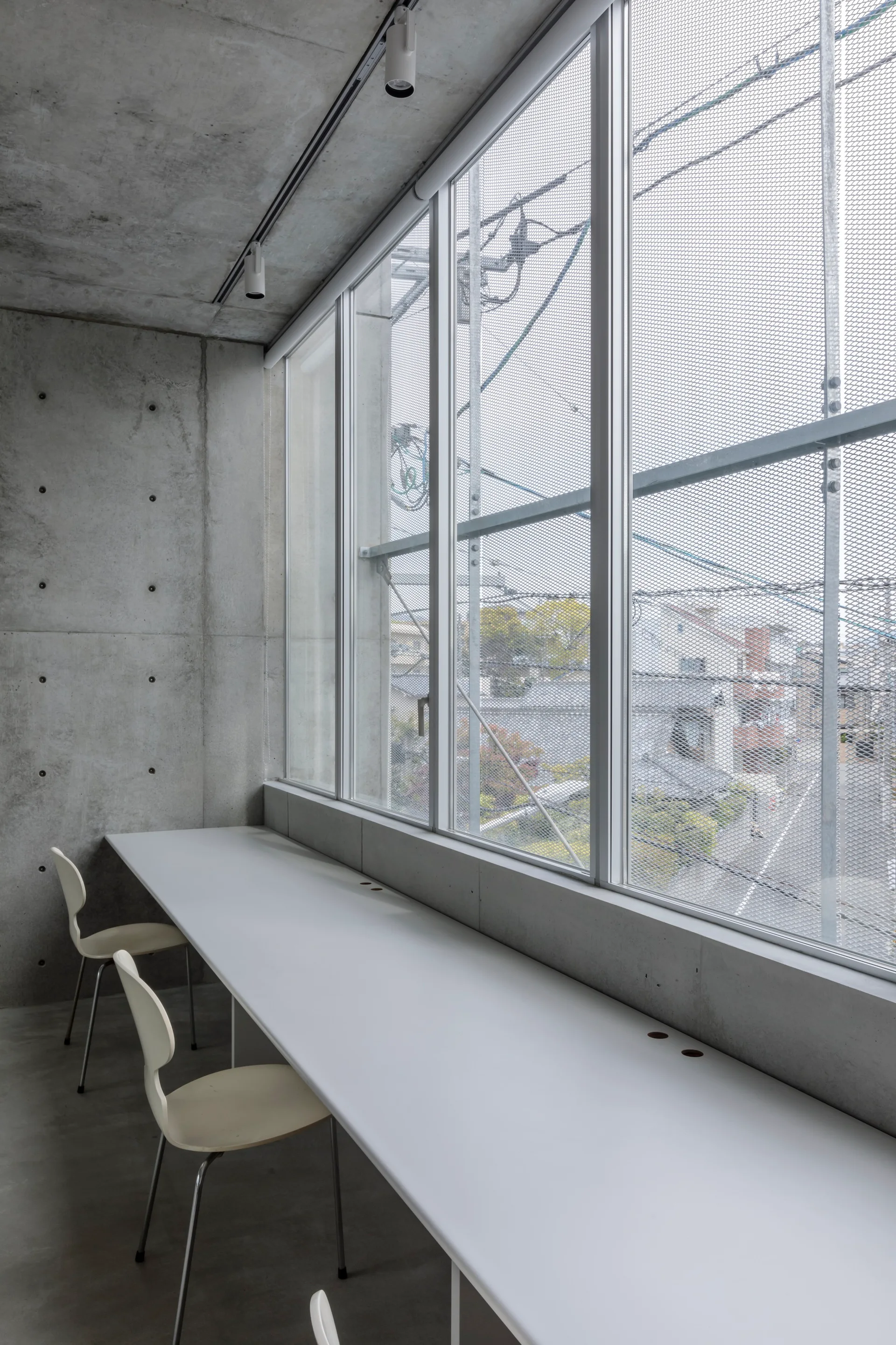 A long white desk and matching minimalist chairs set against raw concrete walls and a large window with a mesh screen.