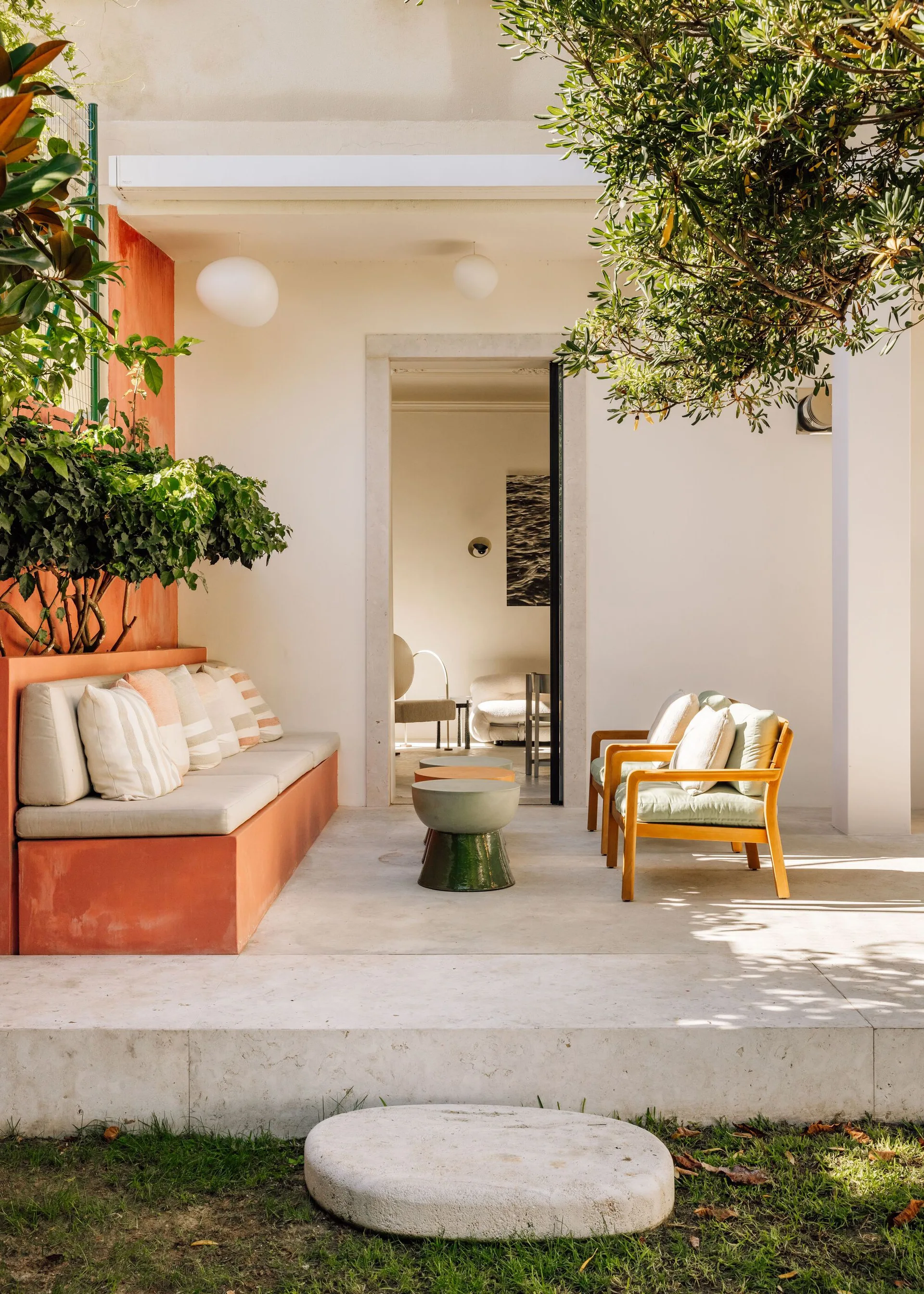 A sunny outdoor patio features a terracotta built-in bench with striped pillows, wooden armchairs, and low nested coffee tables.