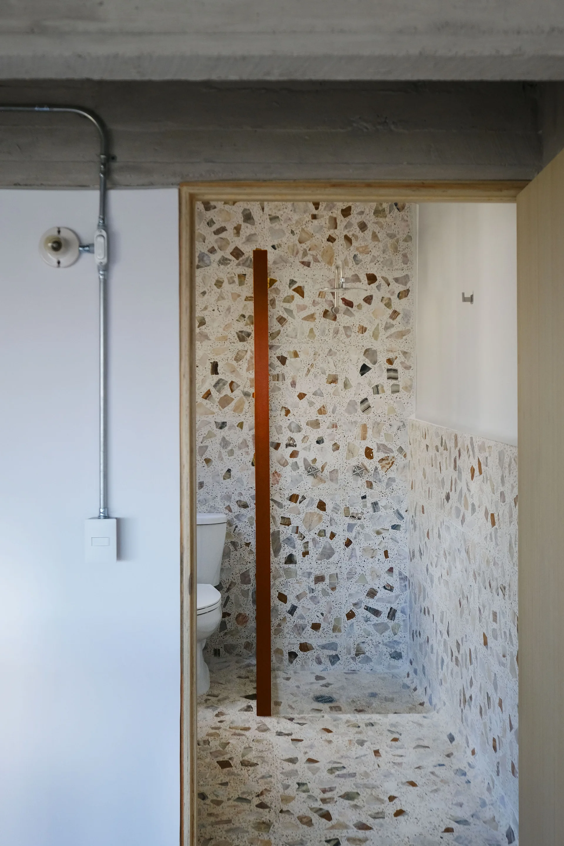 A view into a bathroom featuring large-chip terrazzo tile on the walls and floor, a white toilet, and an exposed concrete ceiling.