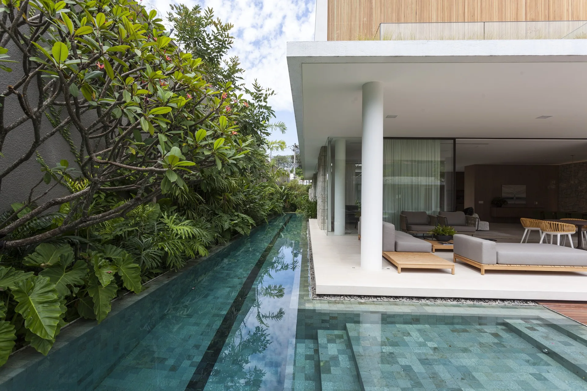 A covered modern terrace with grey lounge furniture sits next to a narrow tiled lap pool bordered by a wall of lush tropical plants.