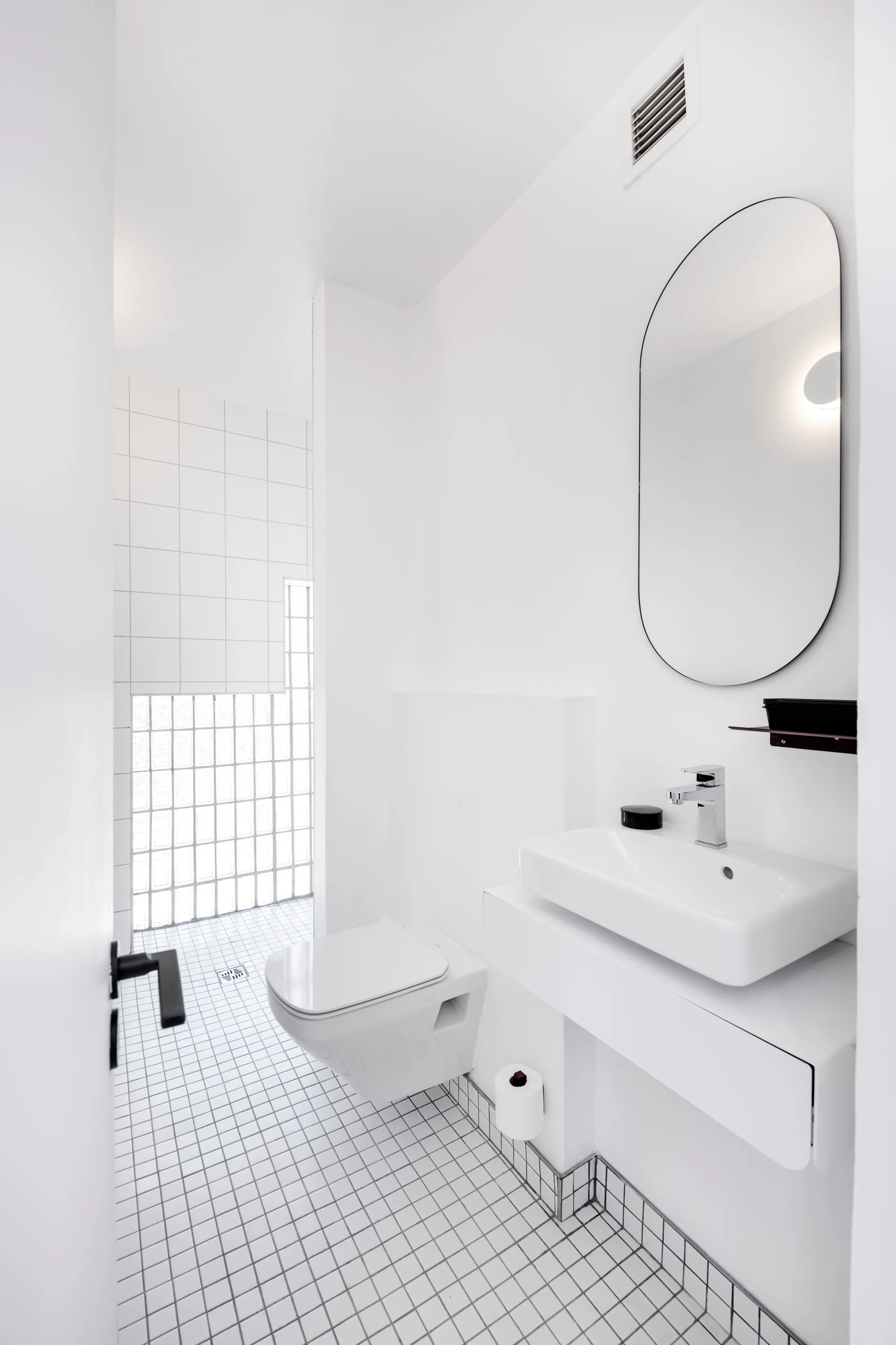 A minimalist white bathroom featuring a wall-mounted sink and toilet, an oval mirror, and small square grid floor tiles near a glass block wall.