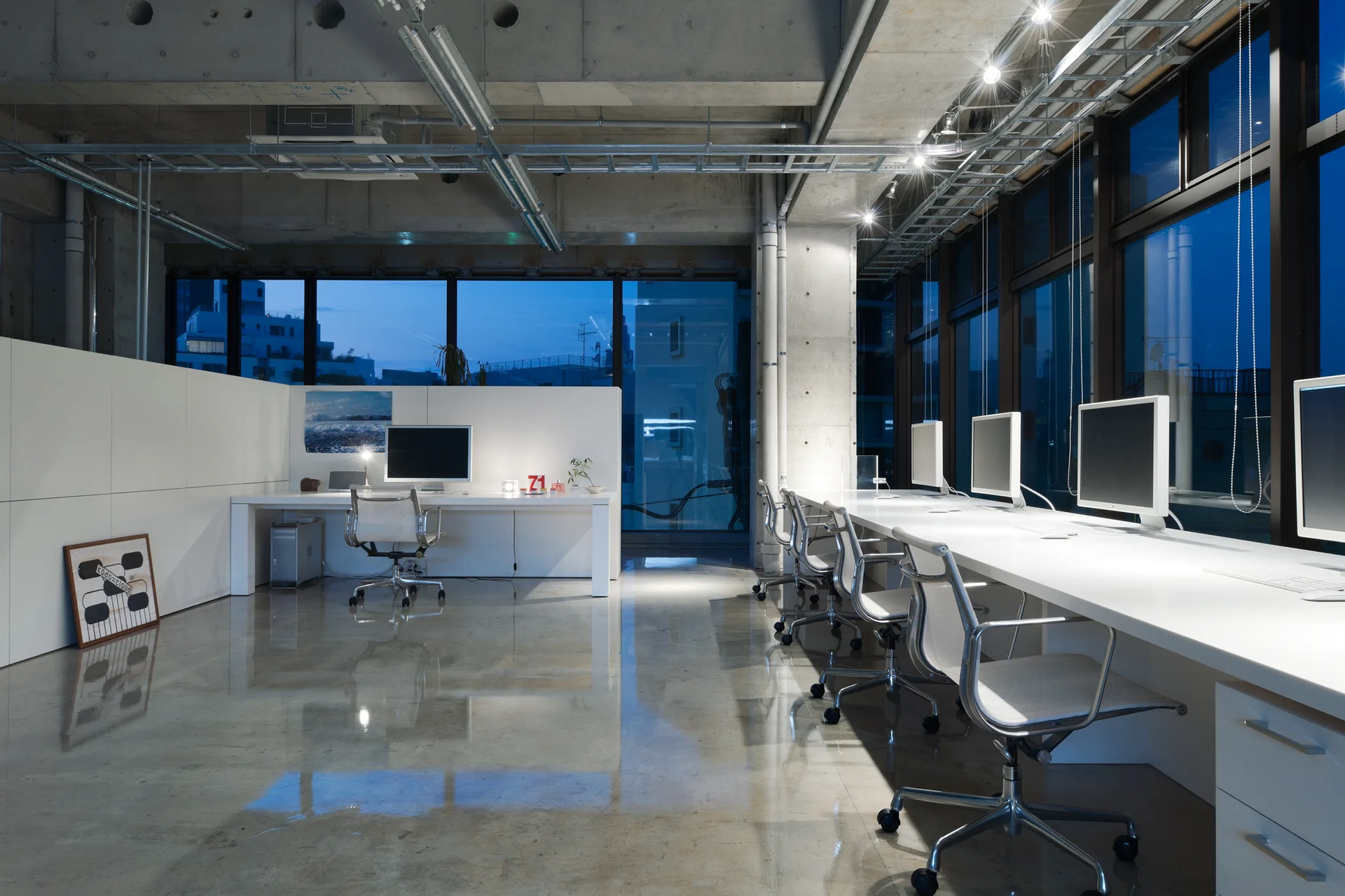 A modern industrial office workspace features white desks, mesh chairs, and large windows set against a polished concrete floor and exposed ceiling.