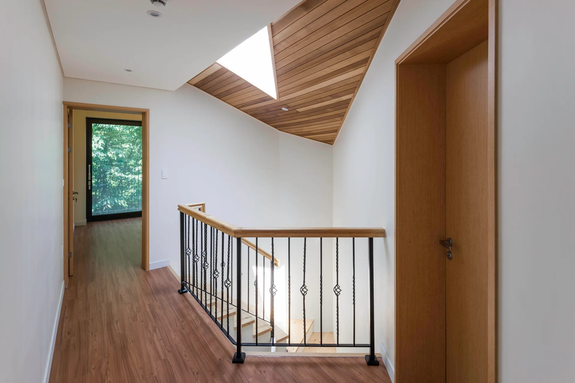 A residential hallway landing featuring a black metal staircase railing, wood flooring, and a slanted wood-plank ceiling with a triangular skylight.