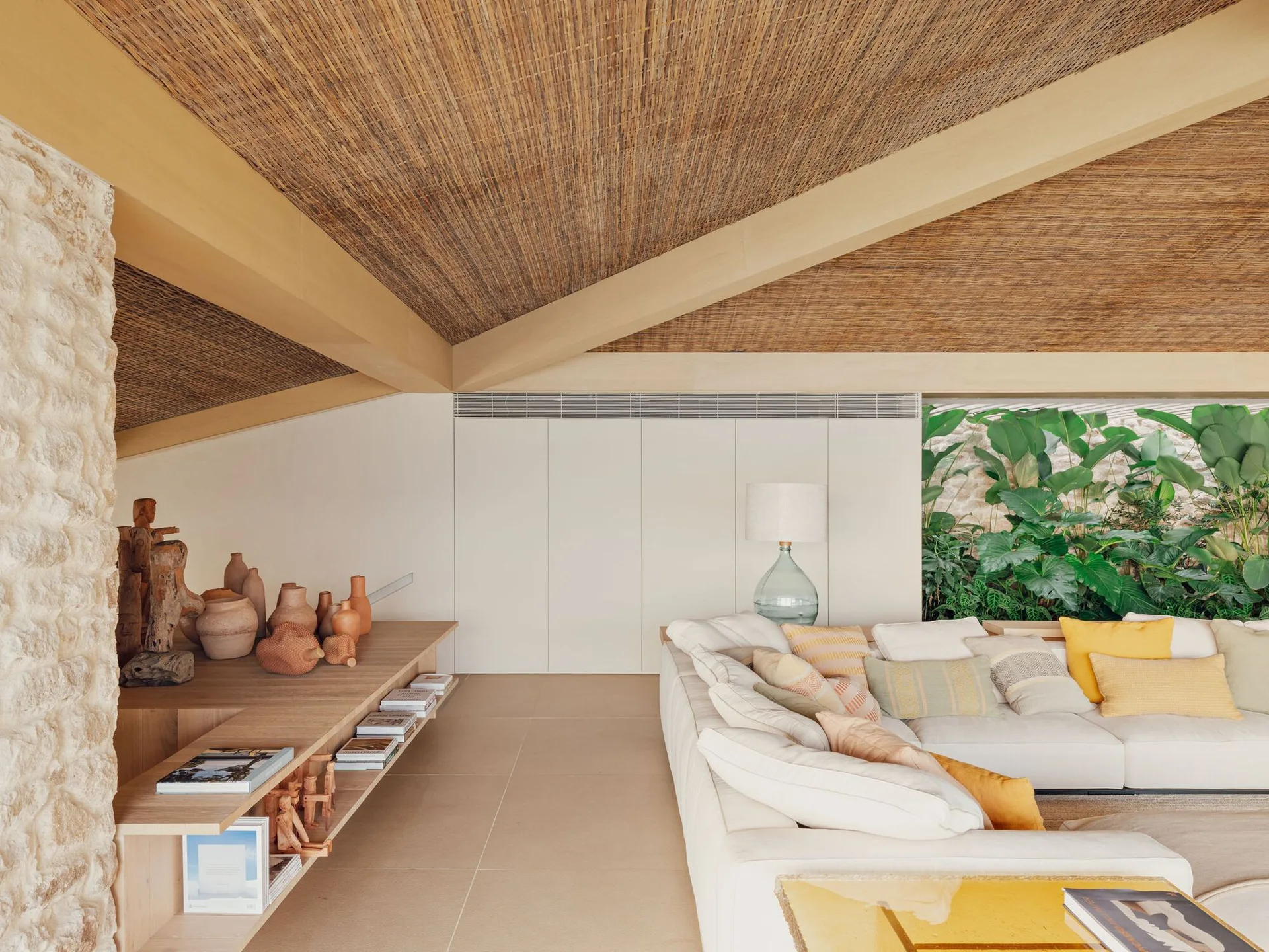 A modern living area featuring a woven reed ceiling with wood beams, a large cream sofa, and a low wood display shelf holding terracotta pottery.