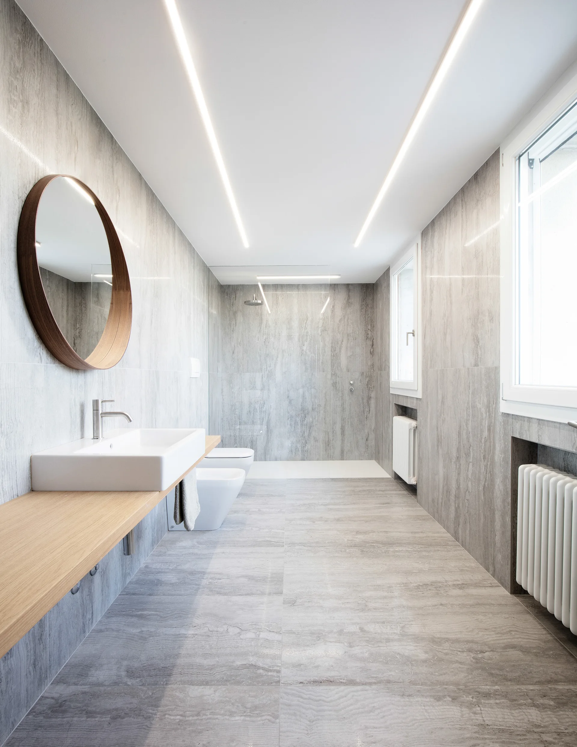 A minimalist bathroom featuring grey stone-like wall and floor tiles, a floating wood vanity with a round mirror, and ceiling linear lighting.